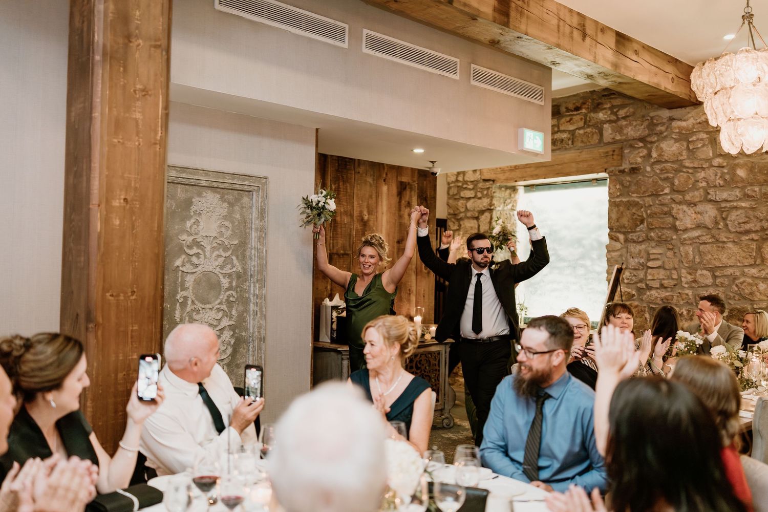 Wedding guests cheering and raising glasses during a reception toast in a rustic stone-walled venue.