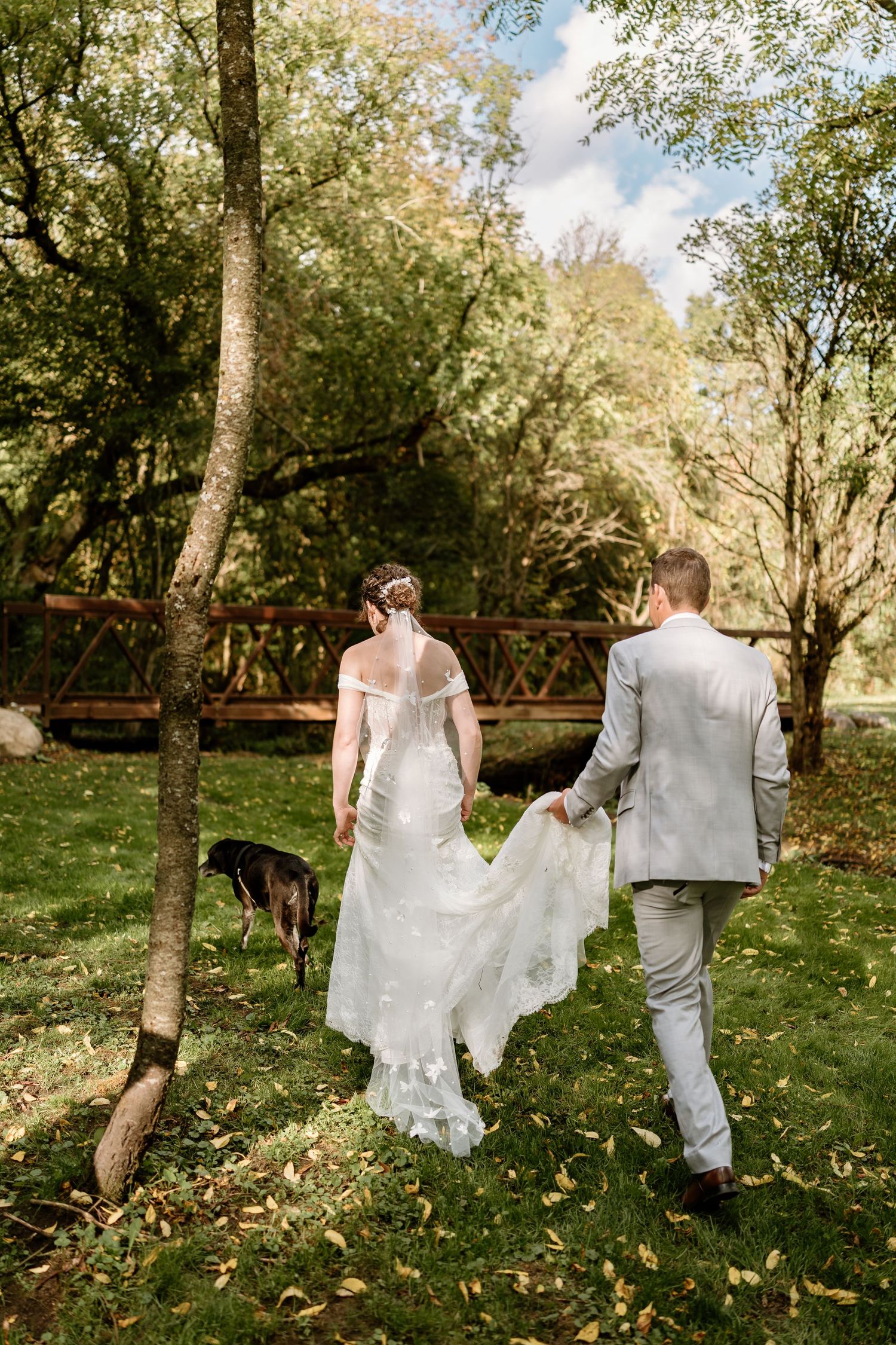 Couple walks through autumn scenery with flowing wedding dress and dog following behind.