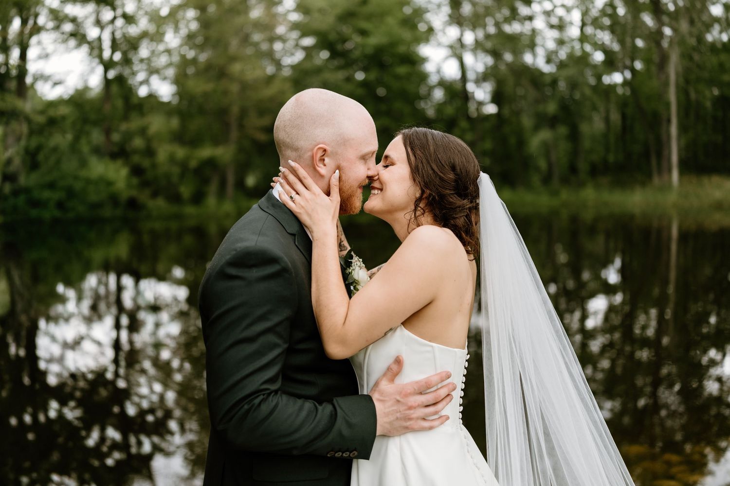 Romantic wedding portrait by lake with trees reflecting in water.