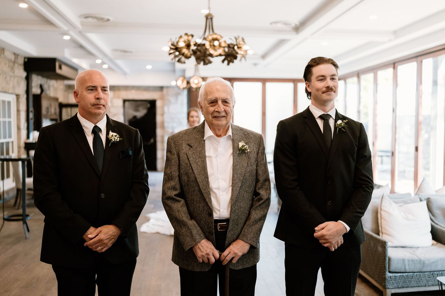 Three men in formal attire stand together in an elegant room with a chandelier.