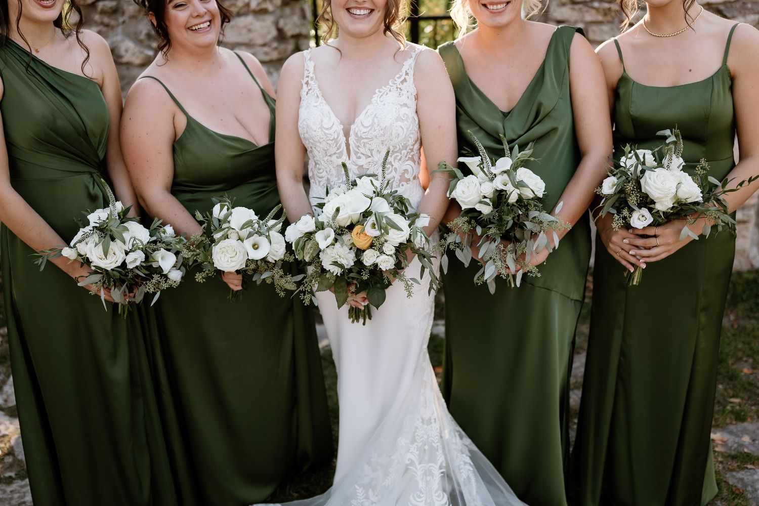 Wedding party members hold white floral bouquets while standing in line against rustic backdrop.