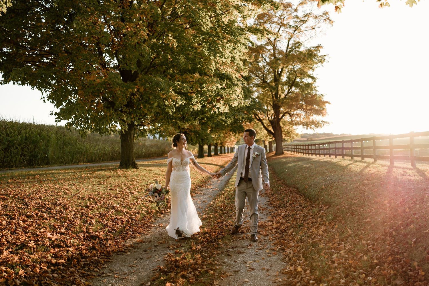 Wedding couple walking along a tree-lined path covered in golden autumn leaves.