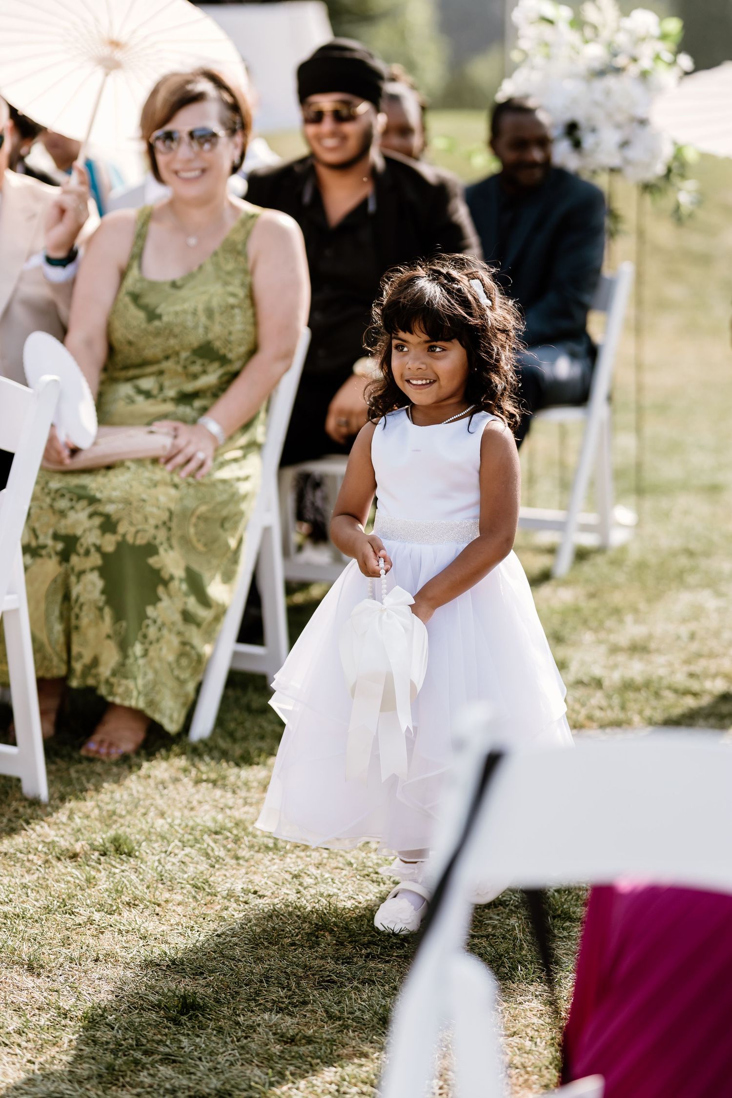 Young flower girl in white dress walks through outdoor wedding ceremony setting.