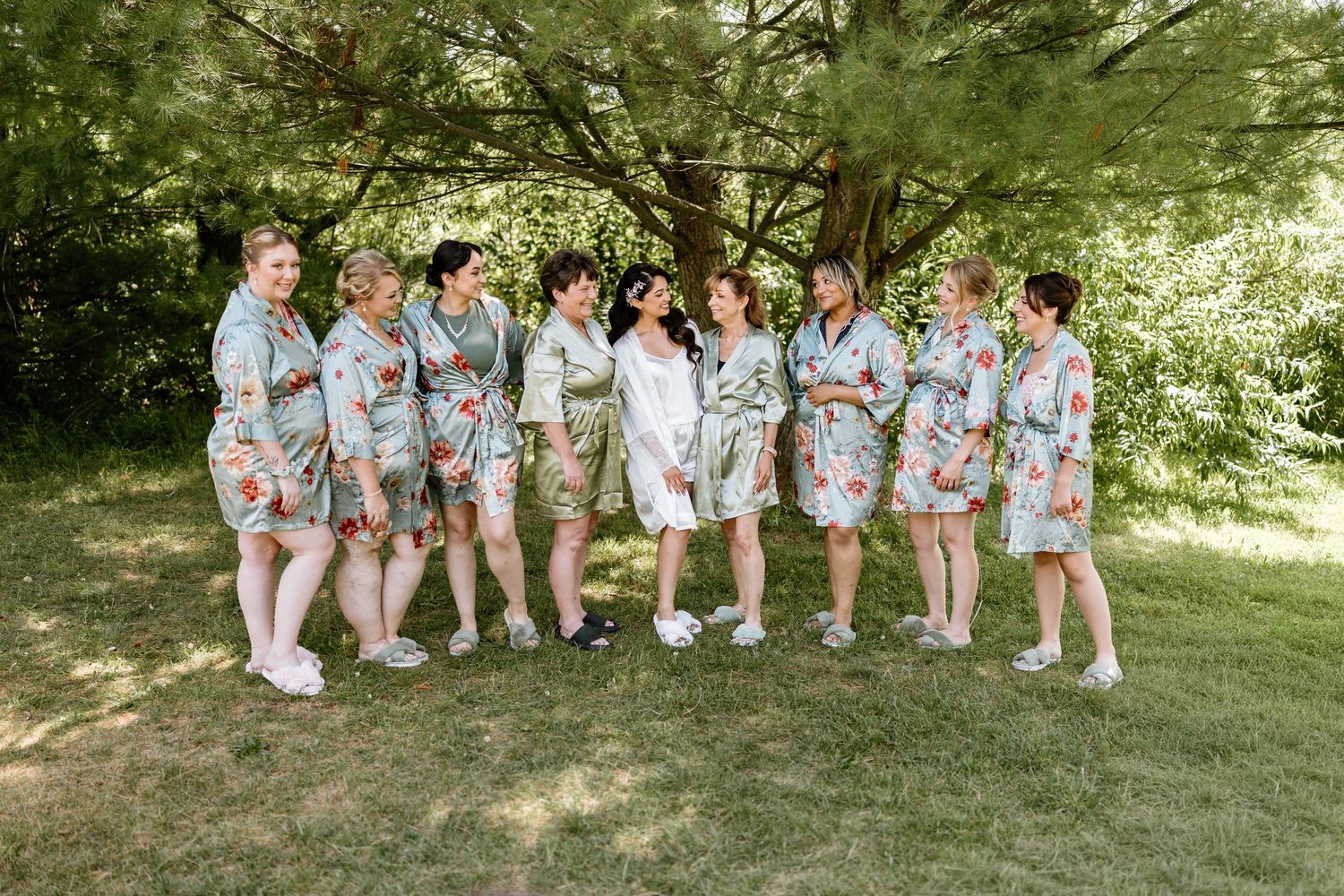 Bridesmaids wearing matching floral robes standing outdoors under green trees before a wedding ceremony.