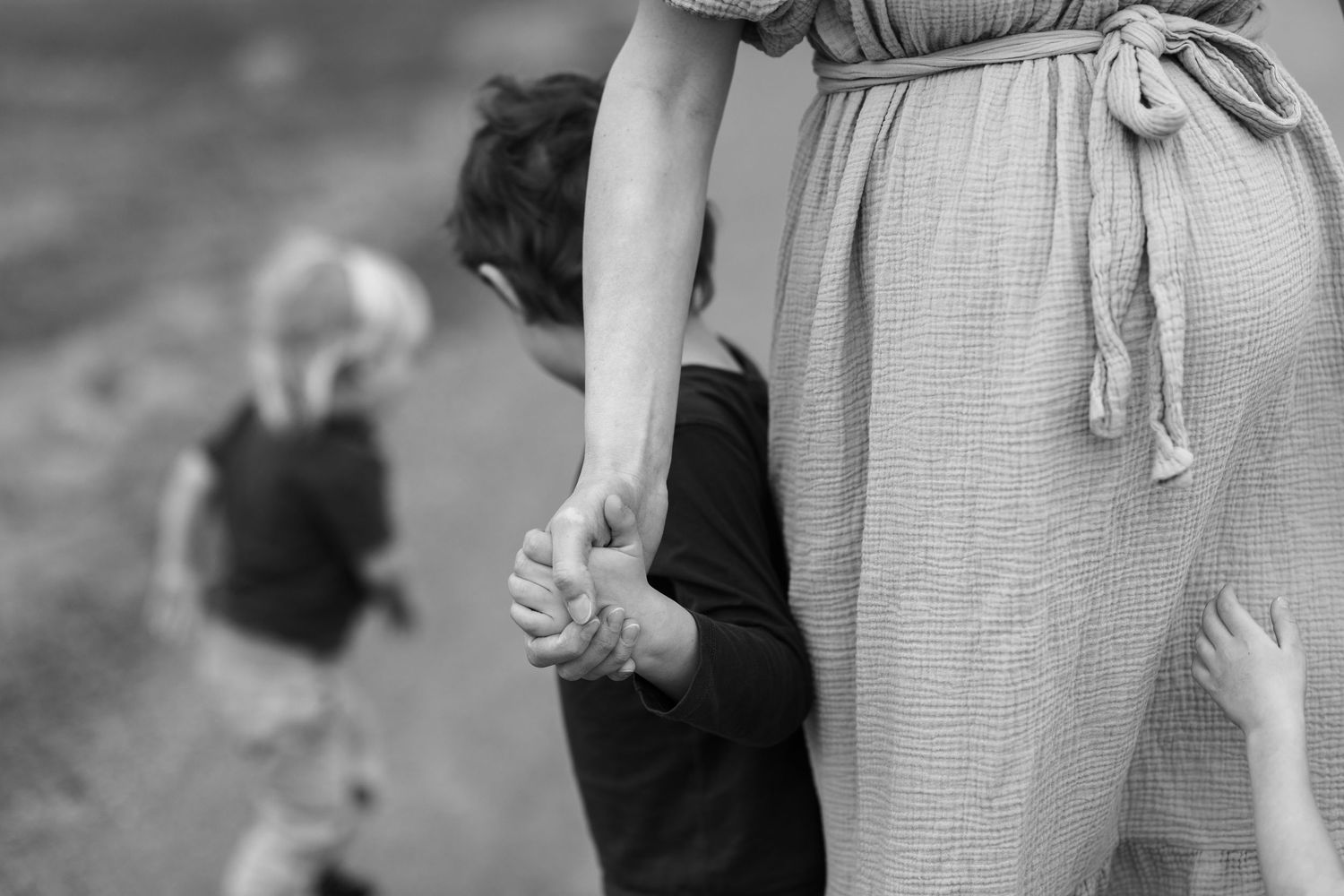 Black and white photo of a pregnant woman holding hands with a child while another toddler walks ahead outdoors.