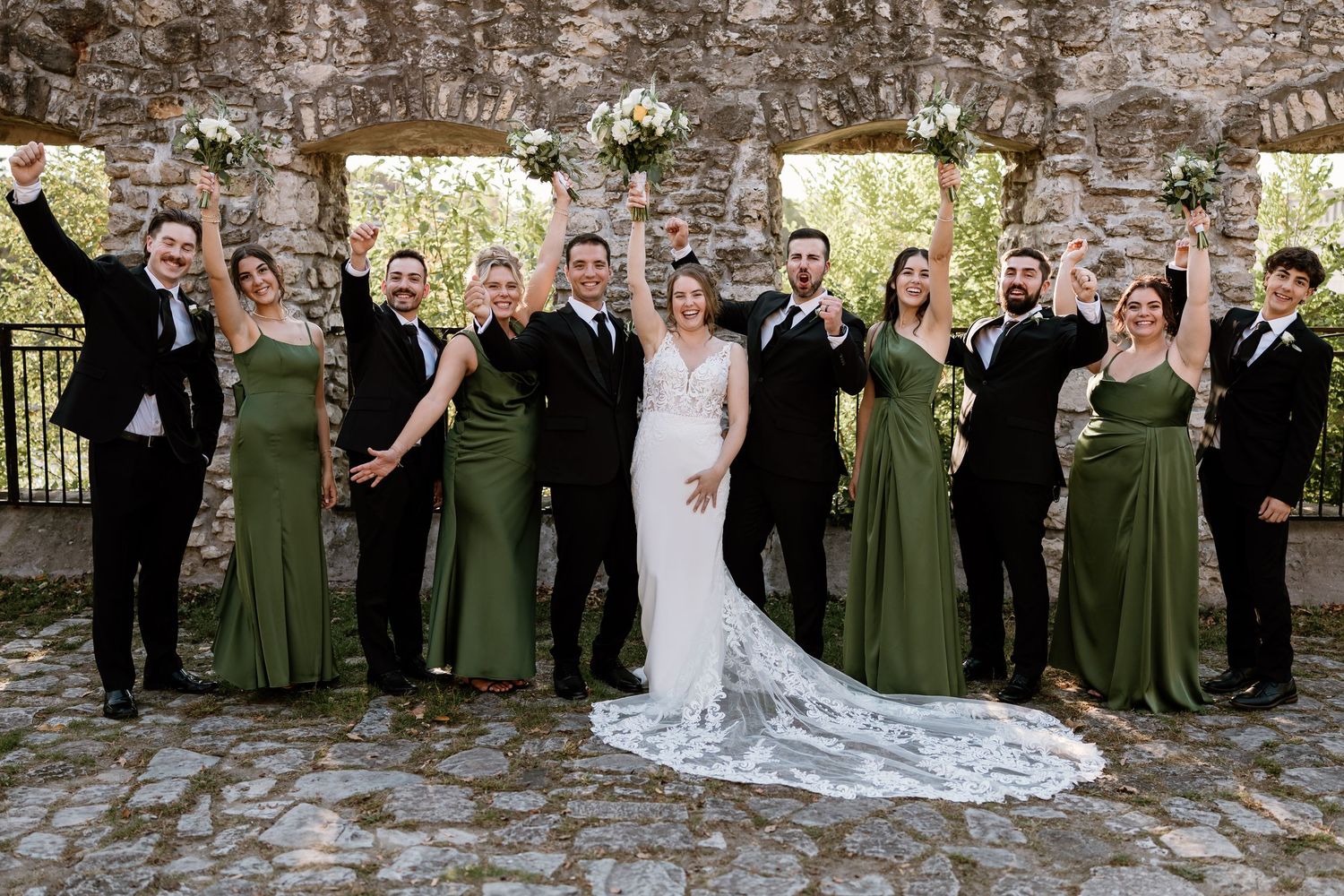 Large wedding party lined up against stone wall with bridesmaids in green and groomsmen in black.