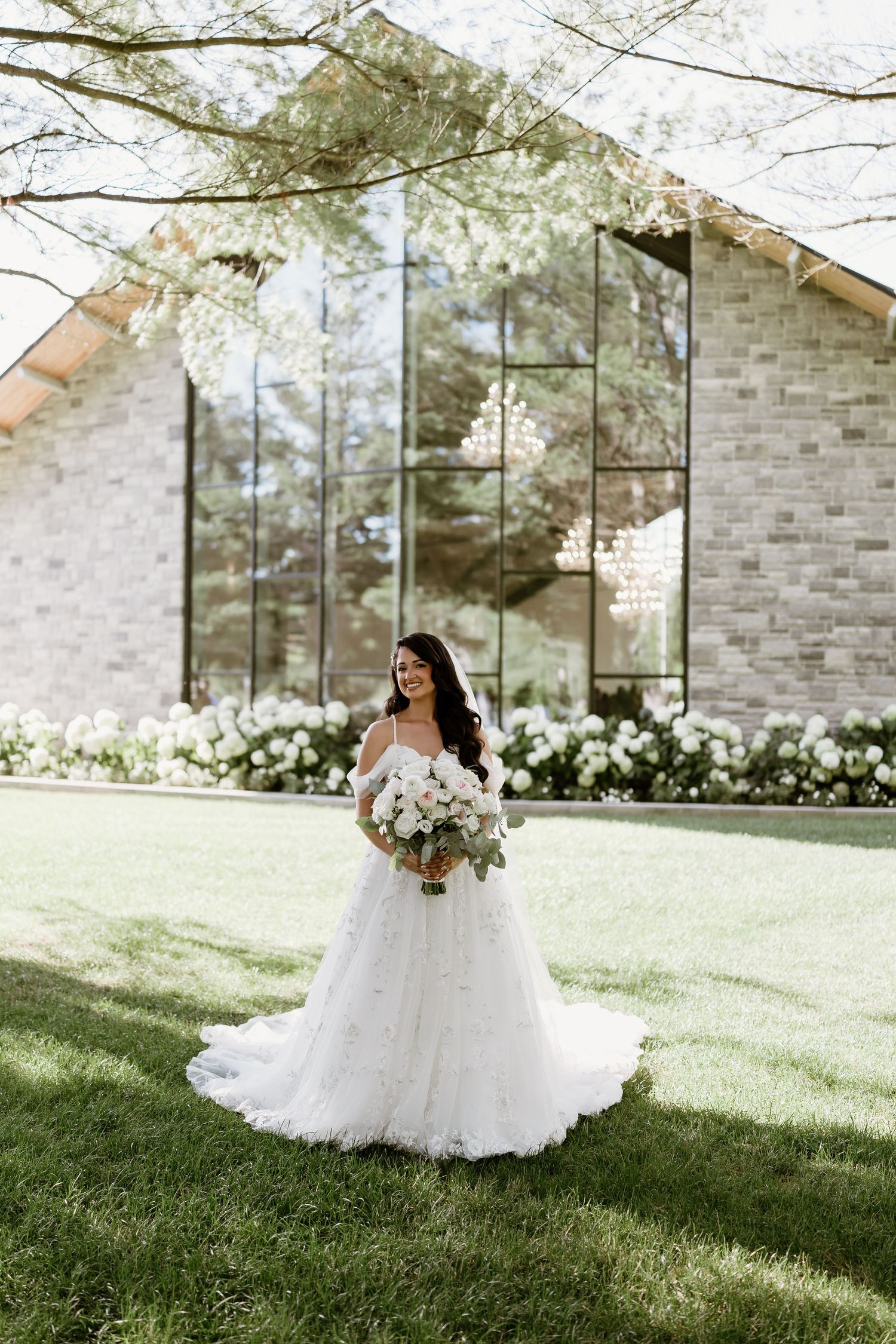 A bride in a flowing white gown poses gracefully against a stone chapel with large windows.
