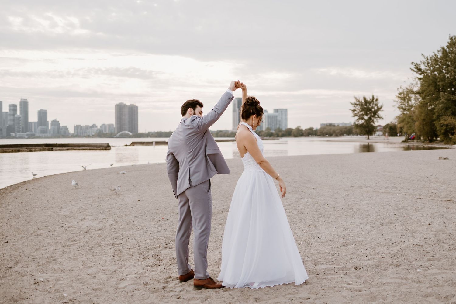 A couple in wedding attire poses for romantic photos on a beach with a city skyline in the background.
