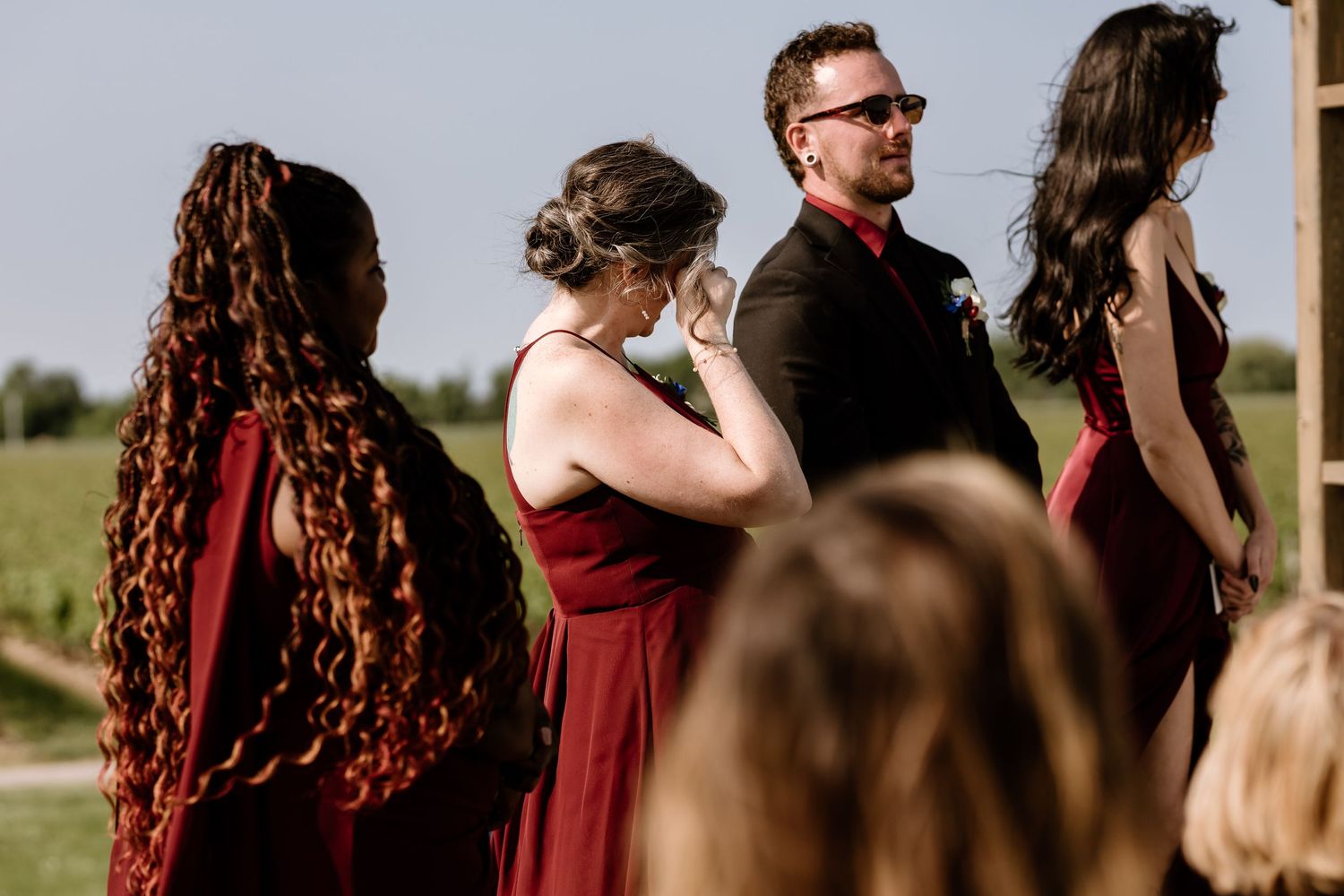 Wedding party members in burgundy dresses standing together during outdoor ceremony.