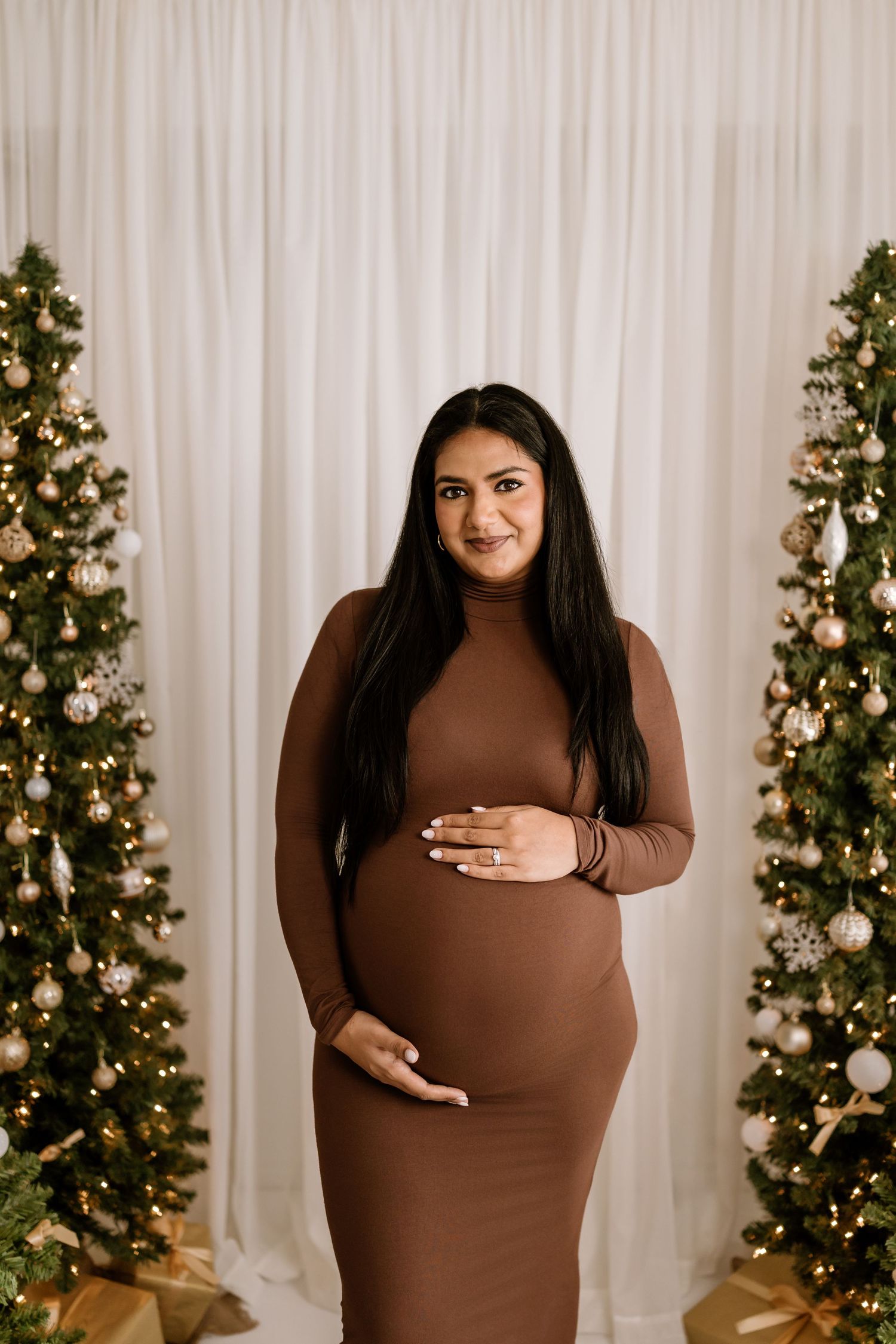 Expectant mother in brown dress poses in front of decorated Christmas trees and wrapped gifts.