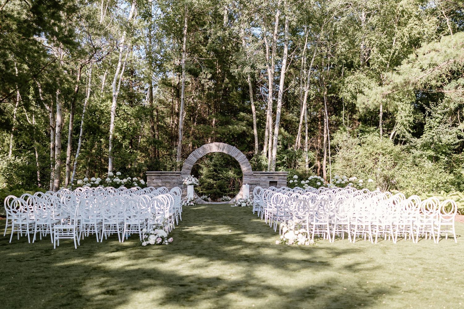 Elegant outdoor wedding ceremony setup with white chairs and floral arch surrounded by tall trees.