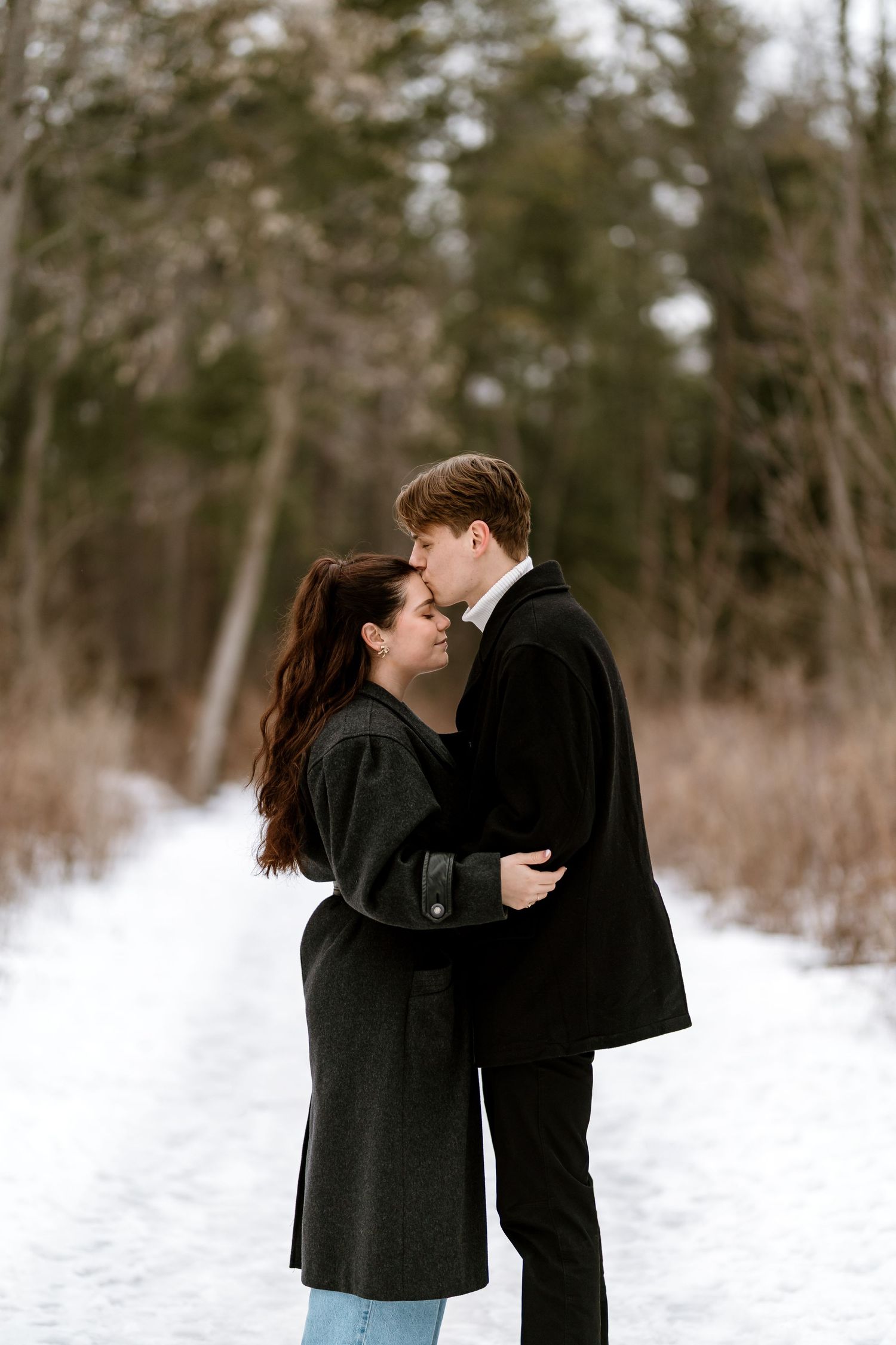A couple shares an intimate moment in a snowy forest setting wearing dark winter coats.