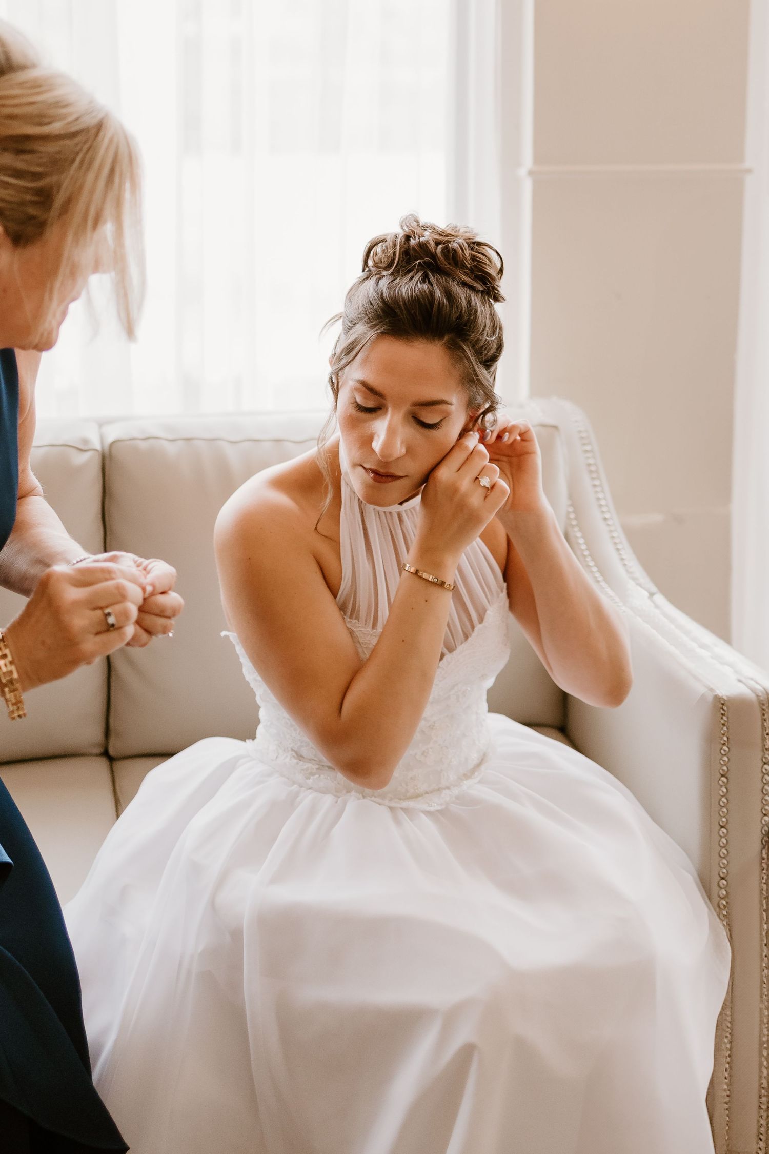 A person in a white wedding gown sits on a beige couch while putting on earrings.