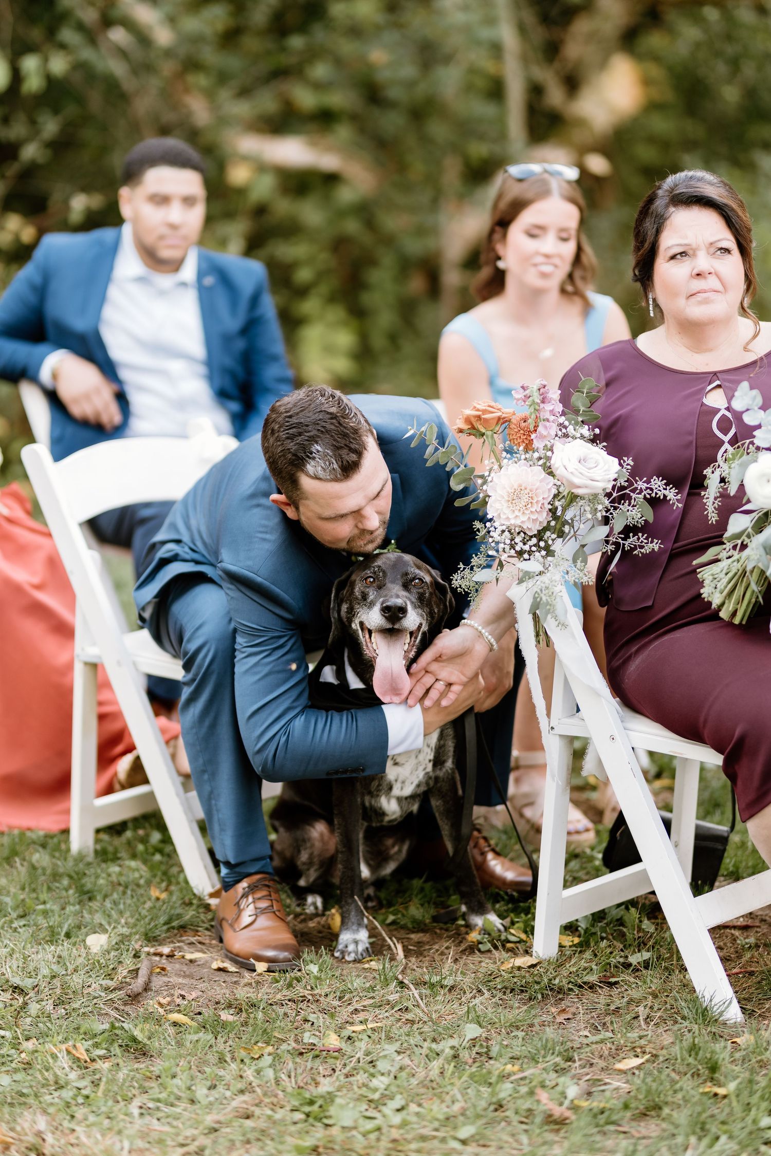 Wedding guest pets black dog during outdoor ceremony while seated on white folding chairs.