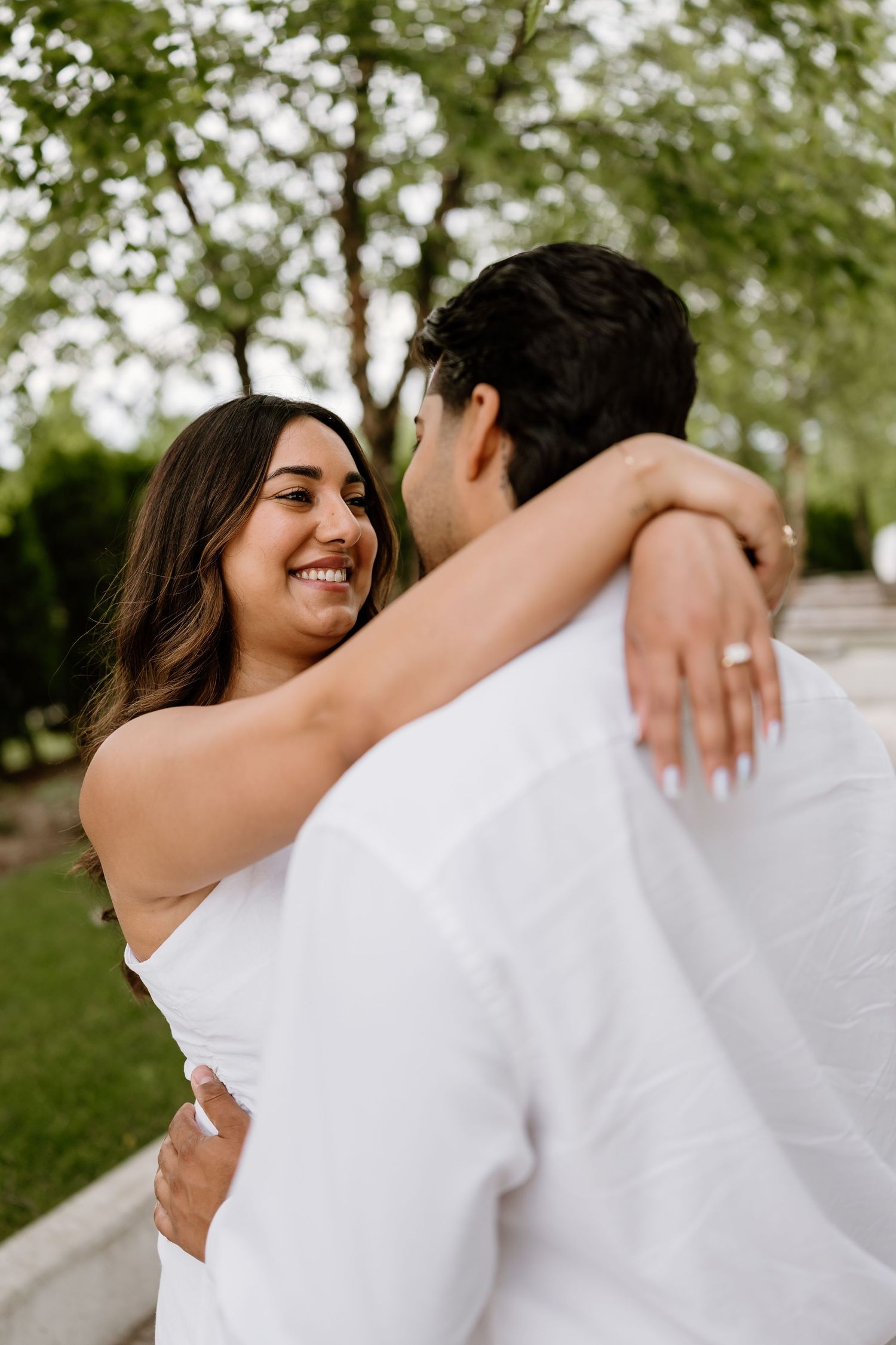A smiling bride in a white dress embraces her groom outdoors, showcasing her engagement ring among lush green trees.
