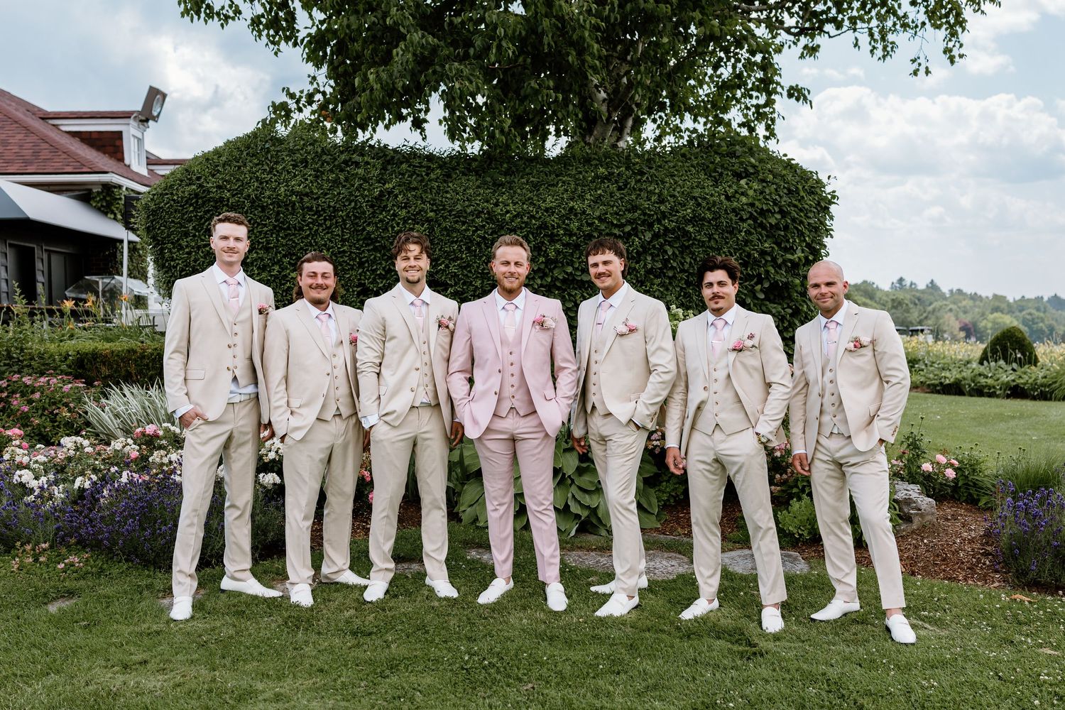 Seven groomsmen in beige suits pose outdoors on green lawn, groom in pink jacket centered, garden wedding photo.