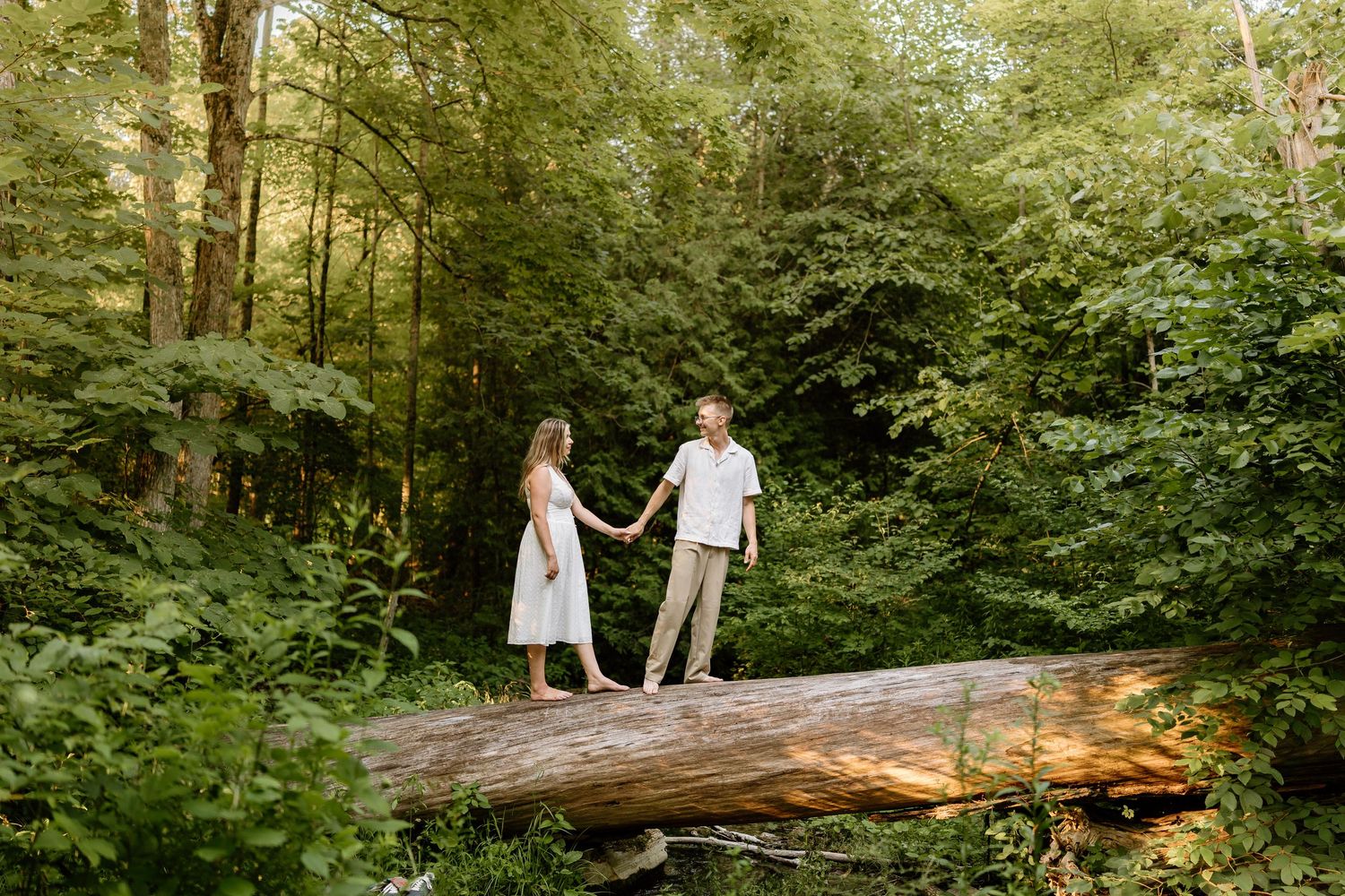 Couple holding hands on a fallen log in a lush green forest, wearing white outfits in a romantic outdoor setting.