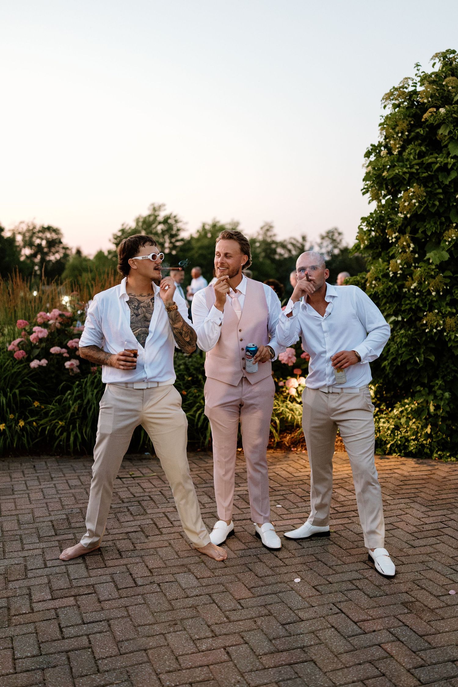 Three men in smart casual attire smile and drink at an outdoor wedding reception surrounded by lush gardens at dusk.