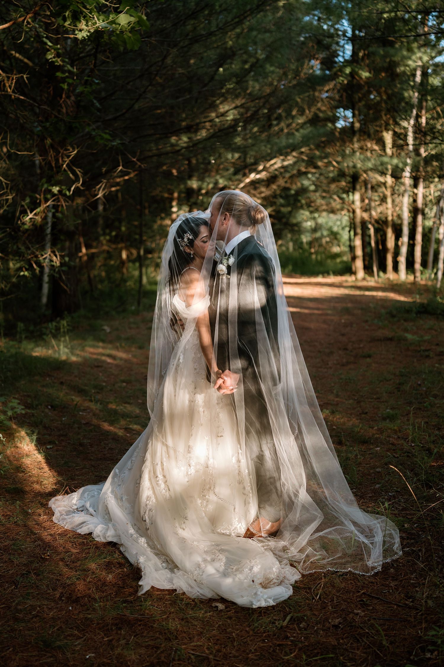 Bride and groom sharing a kiss under a flowing veil in a sunlit forest setting on their wedding day.