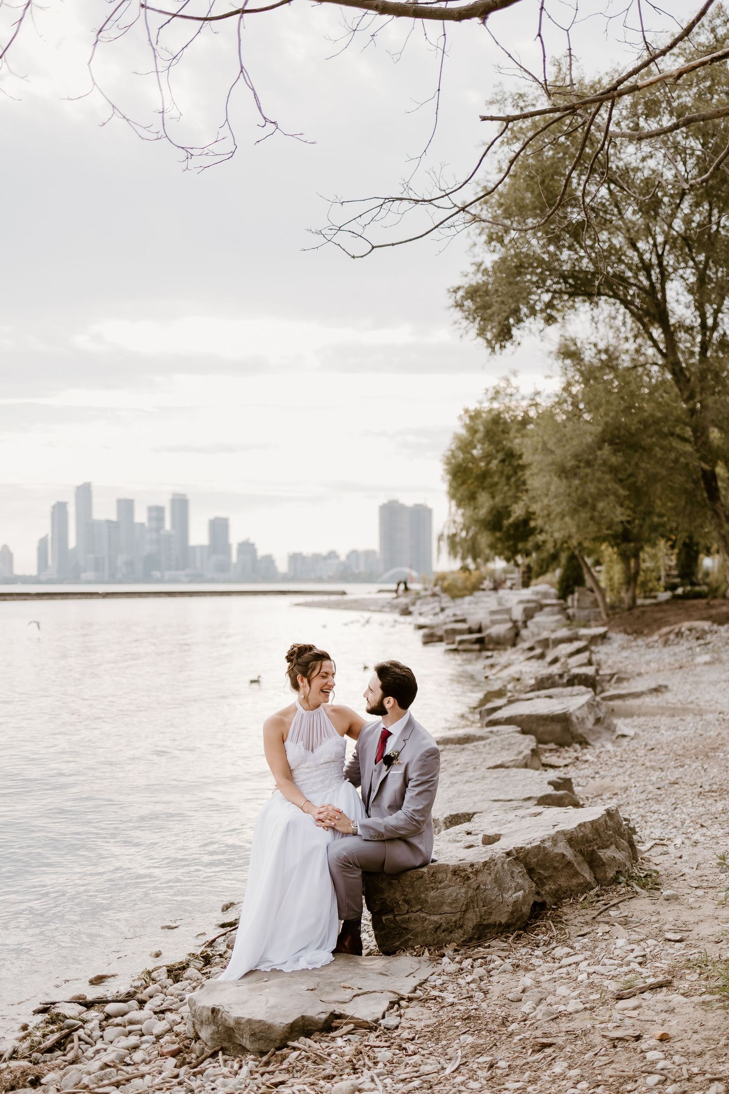 A romantic wedding photoshoot on a rocky shoreline with downtown buildings in the background.