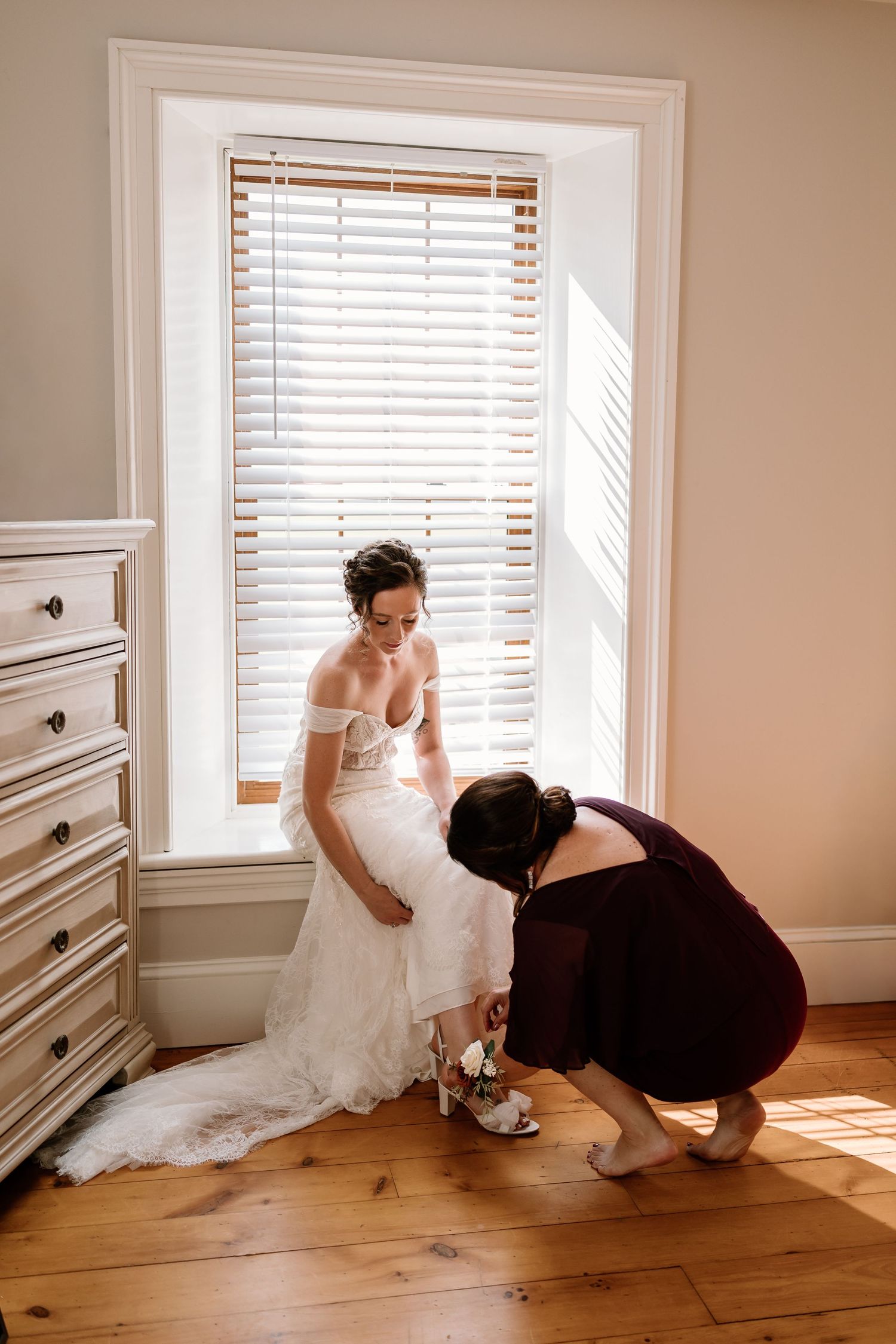 A wedding dress being adjusted in a sunlit room with hardwood floors and white walls.