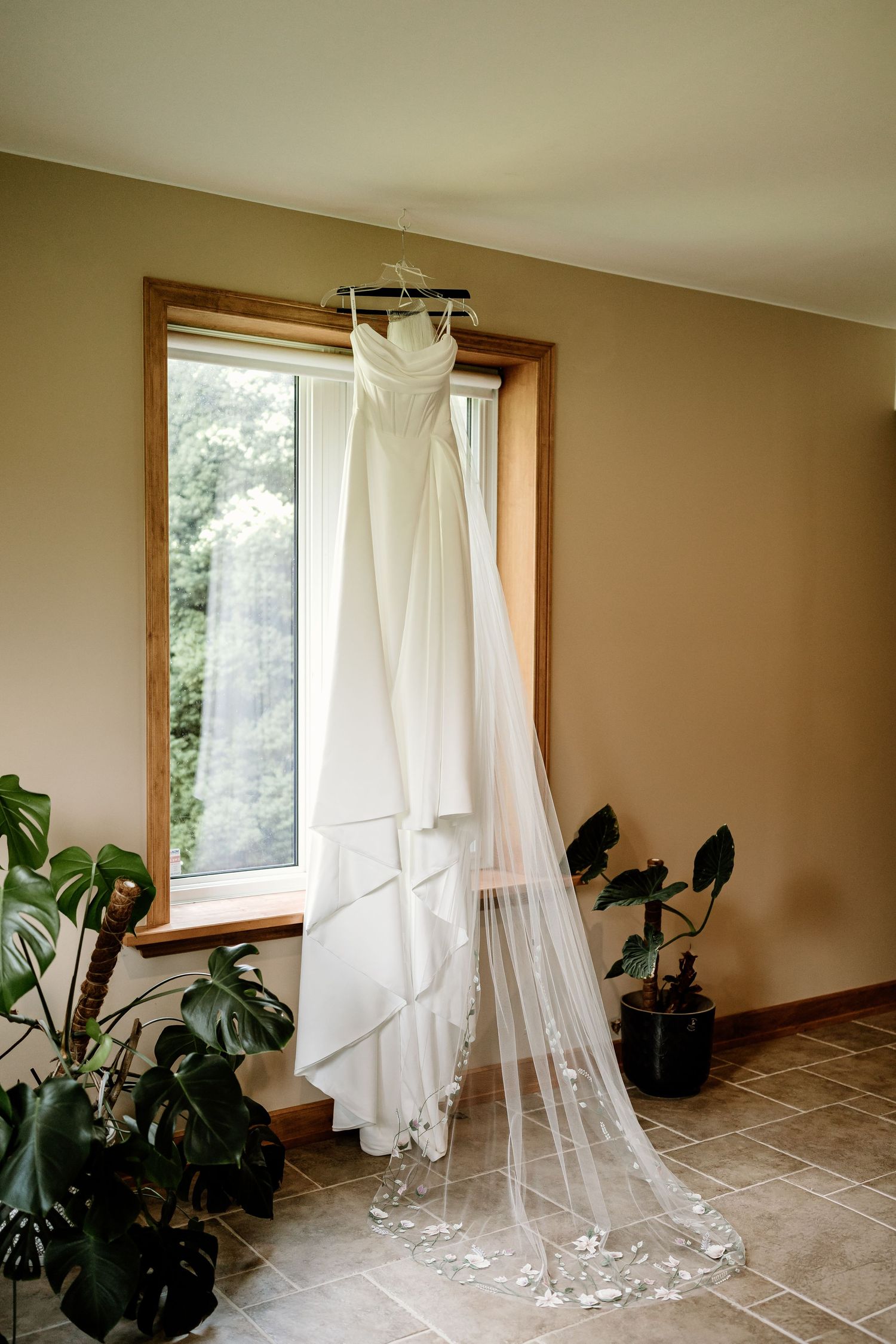 White wedding dress hanging by window with delicate lace details and indoor plants.