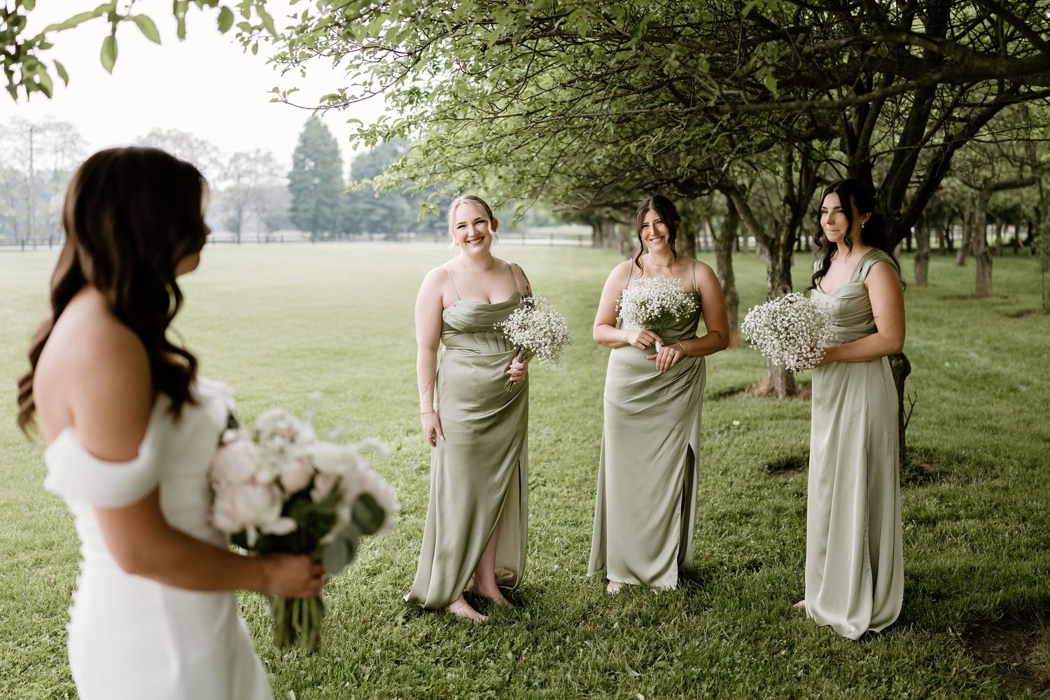 Wedding party in neutral dresses poses on grassy lawn during outdoor summer celebration.