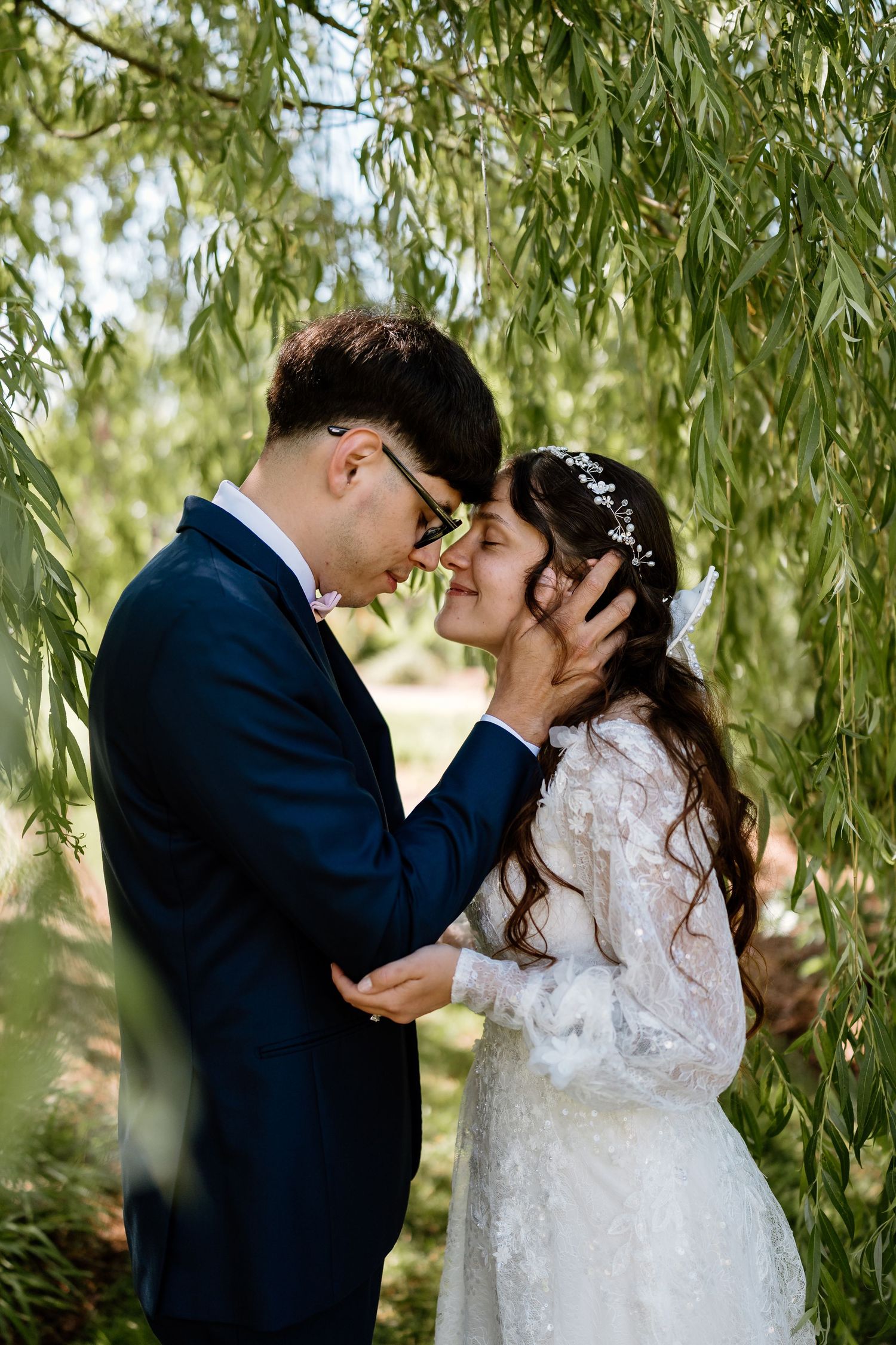 A bride and groom share a romantic moment under a weeping willow tree, with the groom gently holding her face.
