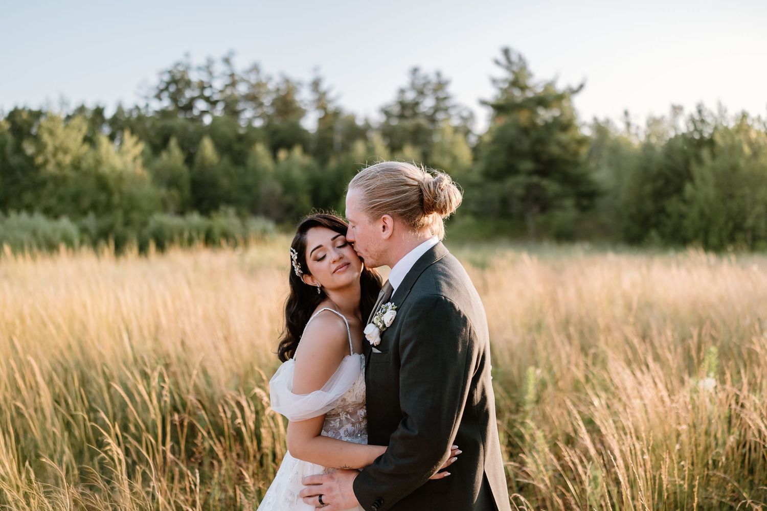 Bride and groom share a romantic embrace in a golden field at sunset on their wedding day.