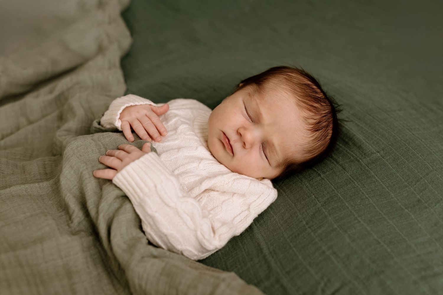Sleeping newborn in light colored outfit resting peacefully on green textured bedding.