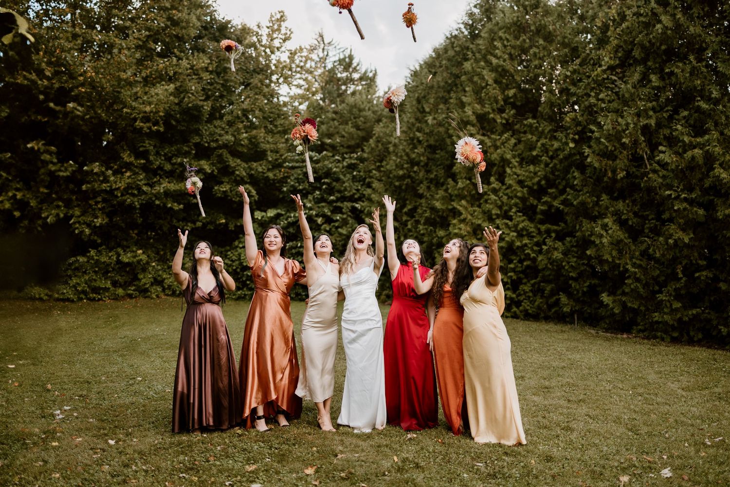 A group of bridesmaids in colorful dresses toss their bouquets in the air during a garden photoshoot.