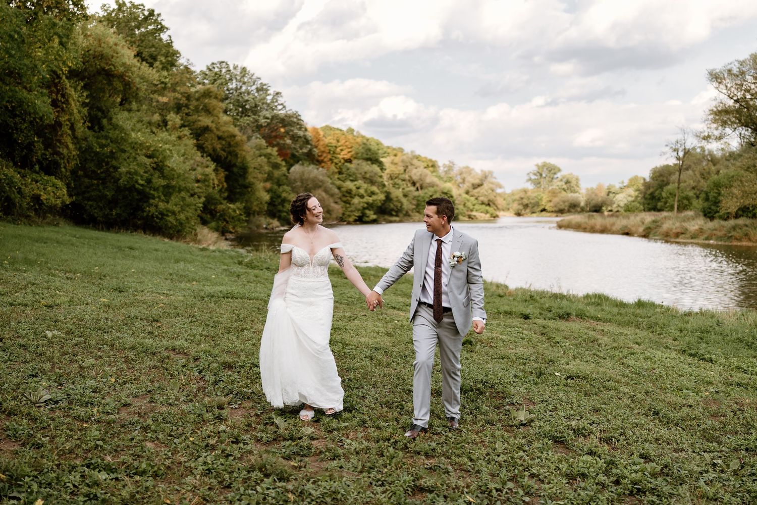 A couple holds hands while walking on grassy riverbank during their wedding day.