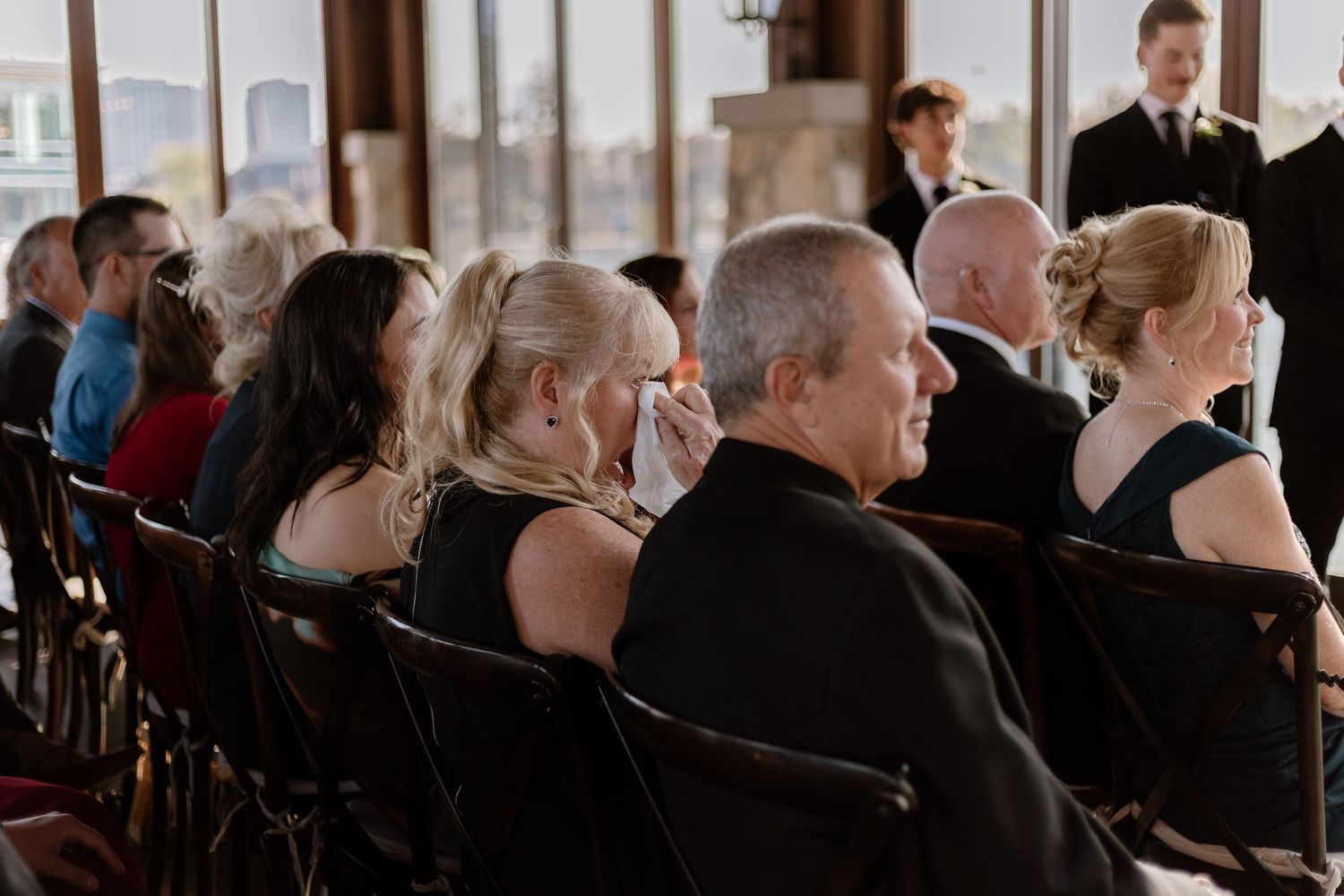 Wedding guests seated in rows watch an indoor ceremony with large windows.