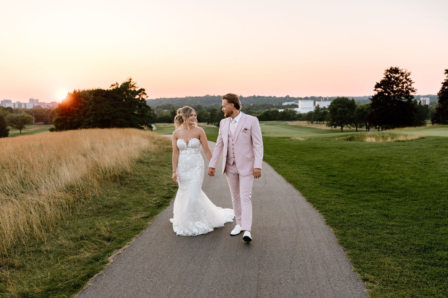 Bride and groom walk hand-in-hand along a golf course path at sunset, she in a mermaid gown, he in a blush suit.