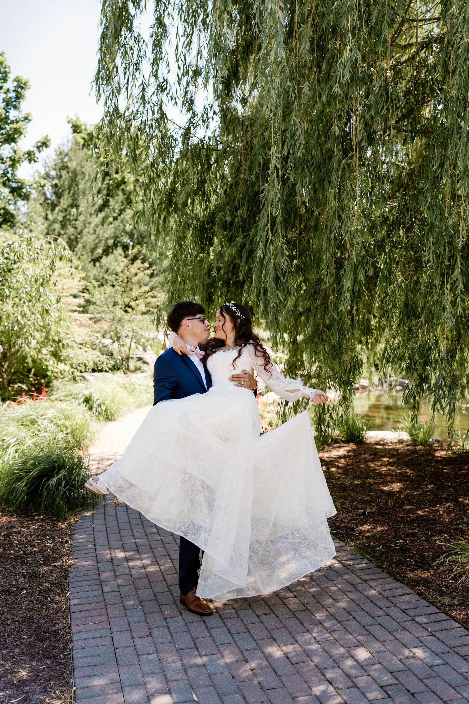 Groom carries bride in white gown under weeping willow tree on brick path in lush garden wedding photo.