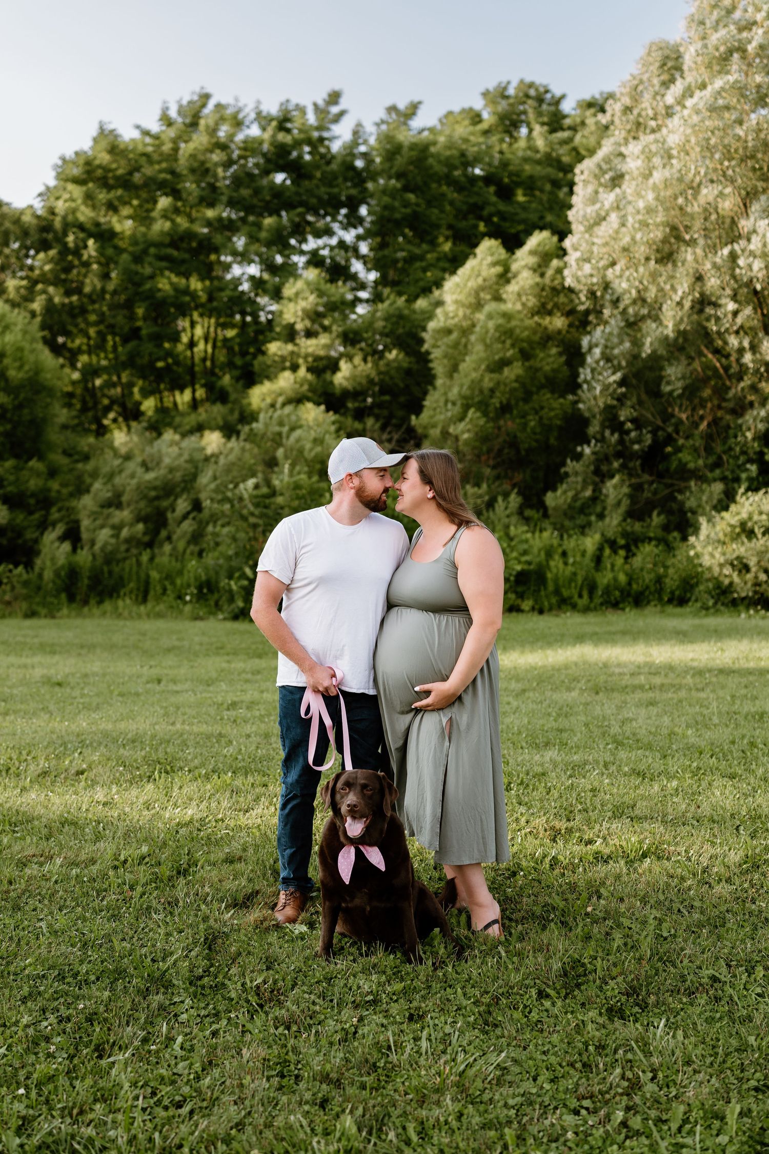Pregnant couple sharing a tender moment in a green park with their chocolate Labrador wearing a pink bow leash.