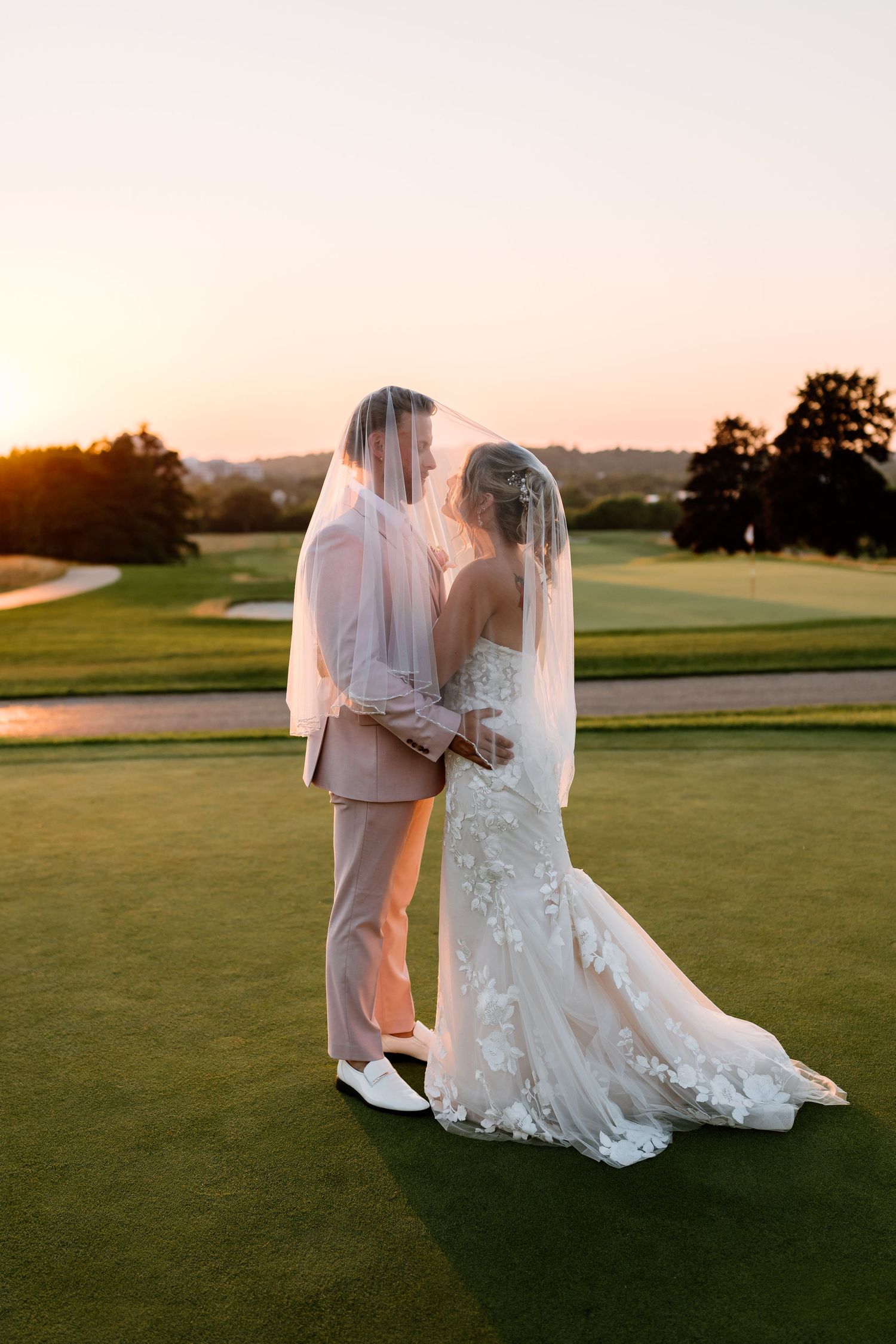 Bride and groom share a romantic moment under a veil at sunset on a lush green golf course.