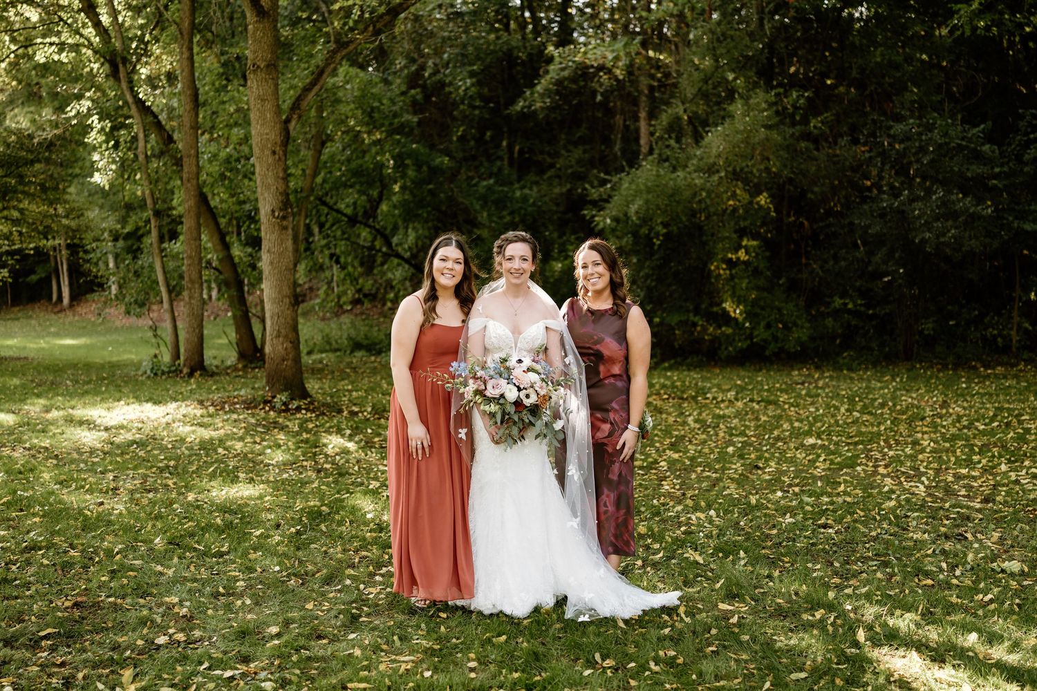 Bride in white gown poses with bridesmaids wearing rust and burgundy dresses on a fall day.