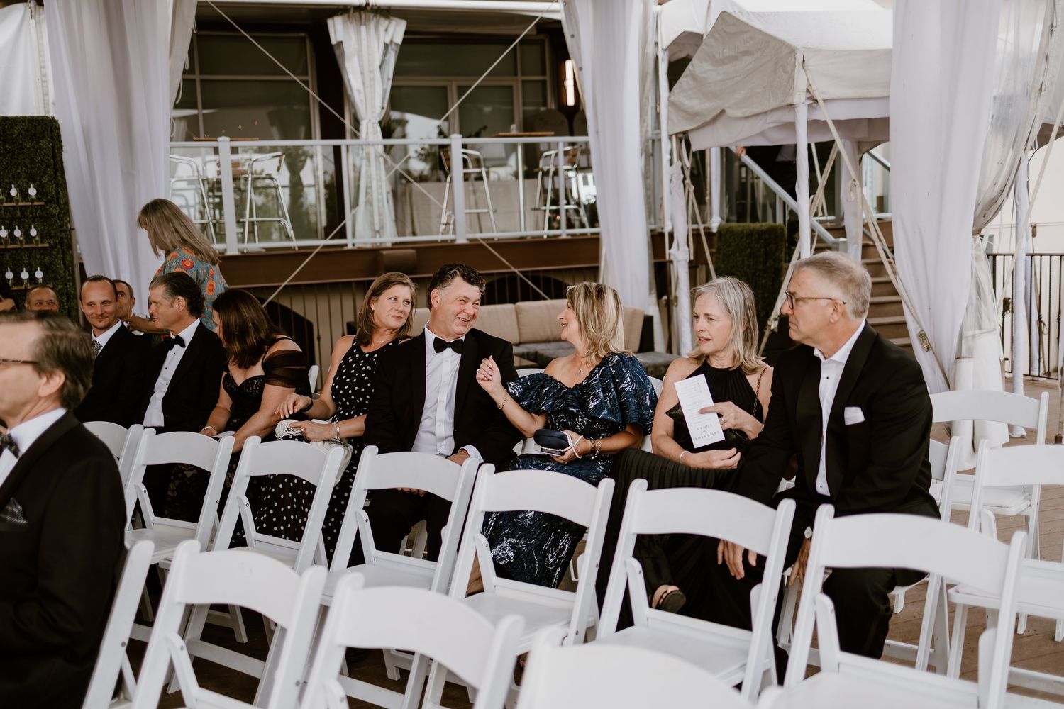 Wedding guests seated in white chairs under an elegant white tent structure.