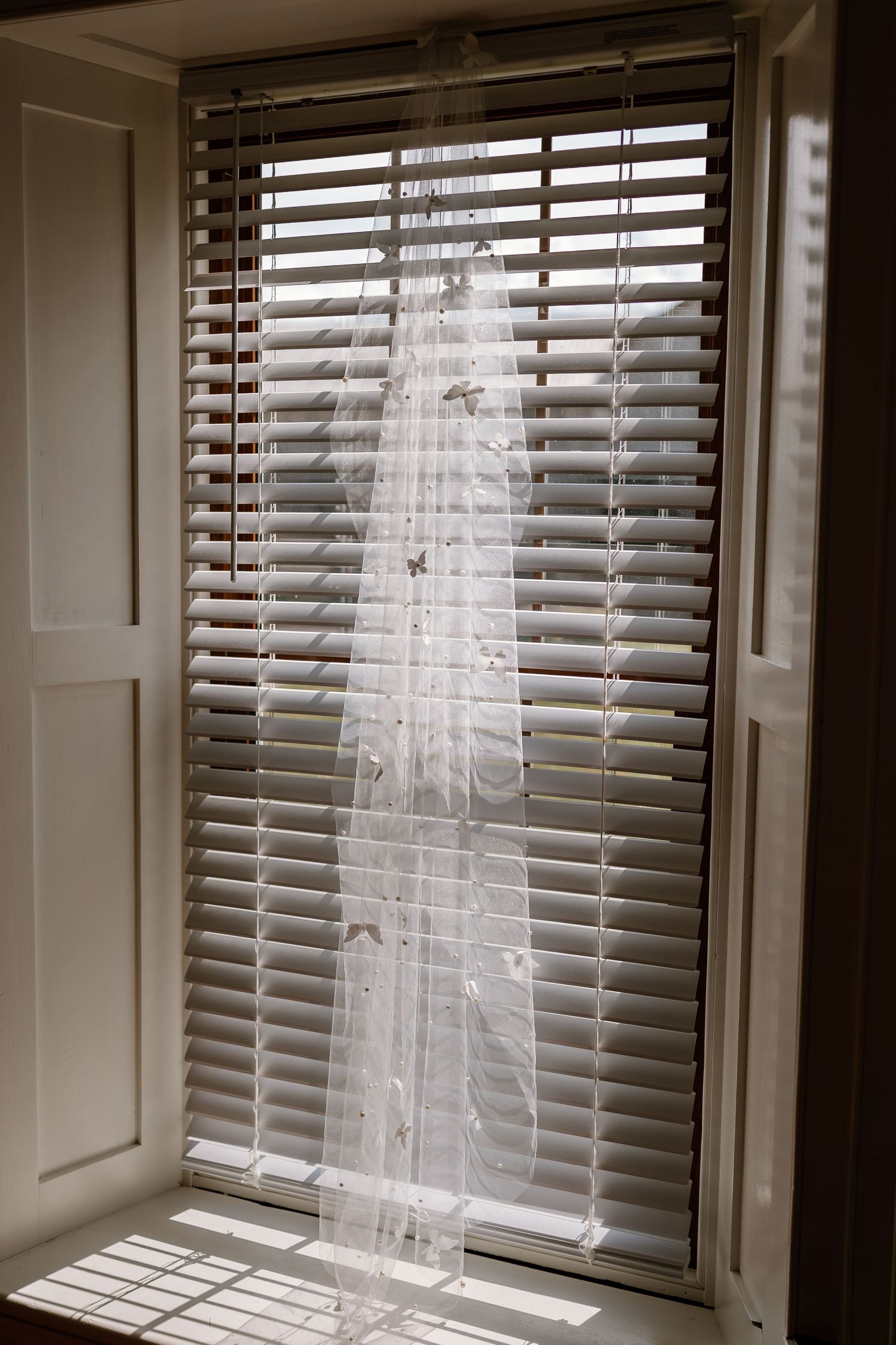 White sheer curtain hanging against venetian blinds creating a soft, ethereal effect.
