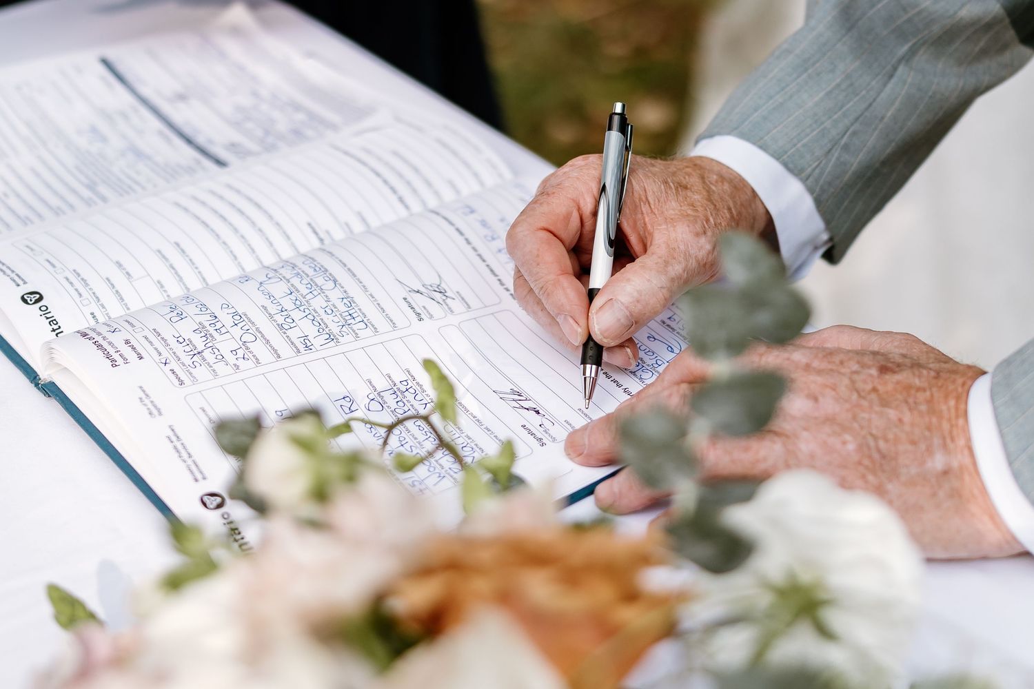 Close-up of hands signing a wedding document with white flowers in the foreground.