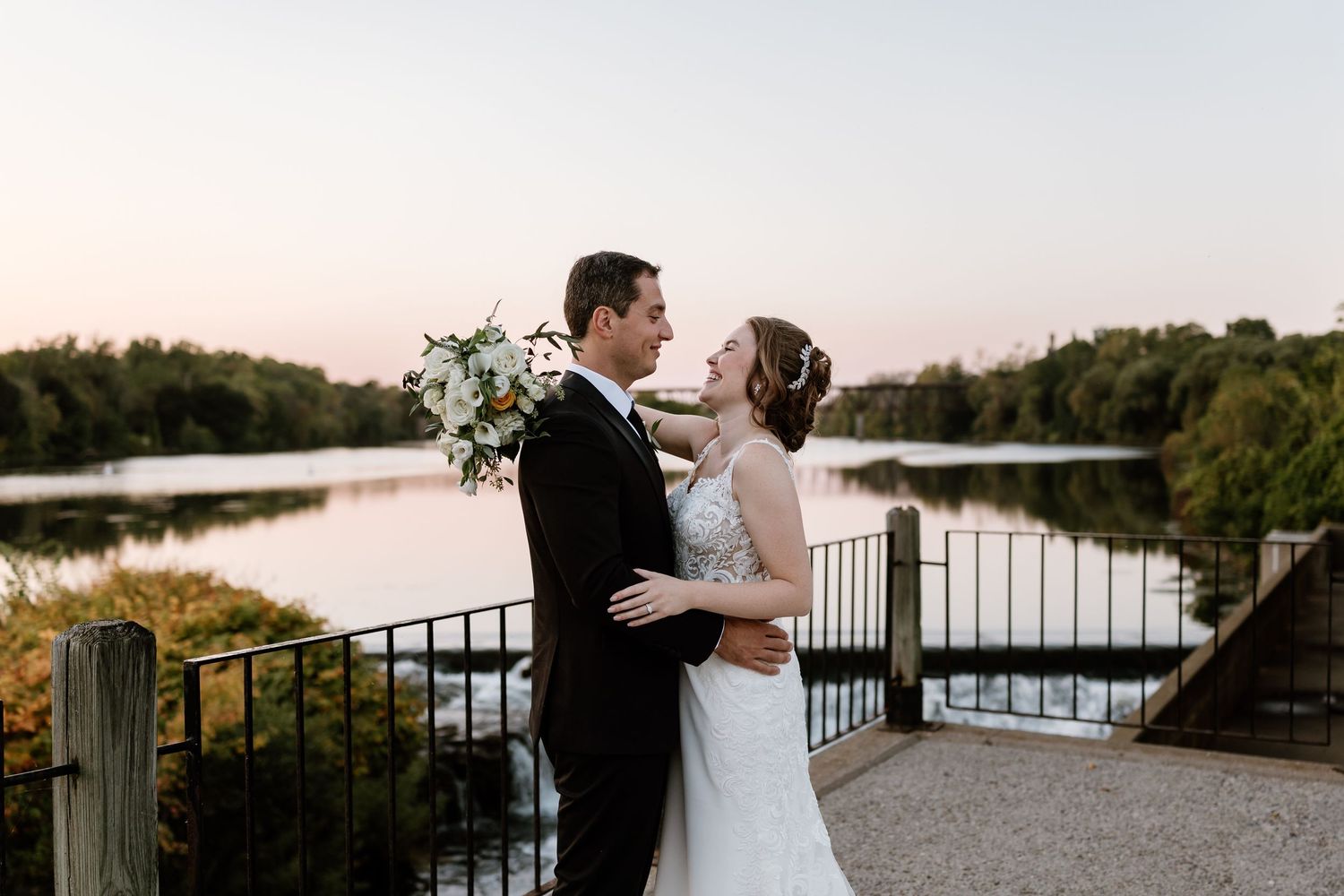 A romantic sunset wedding portrait of a couple embracing by a lakeside railing.