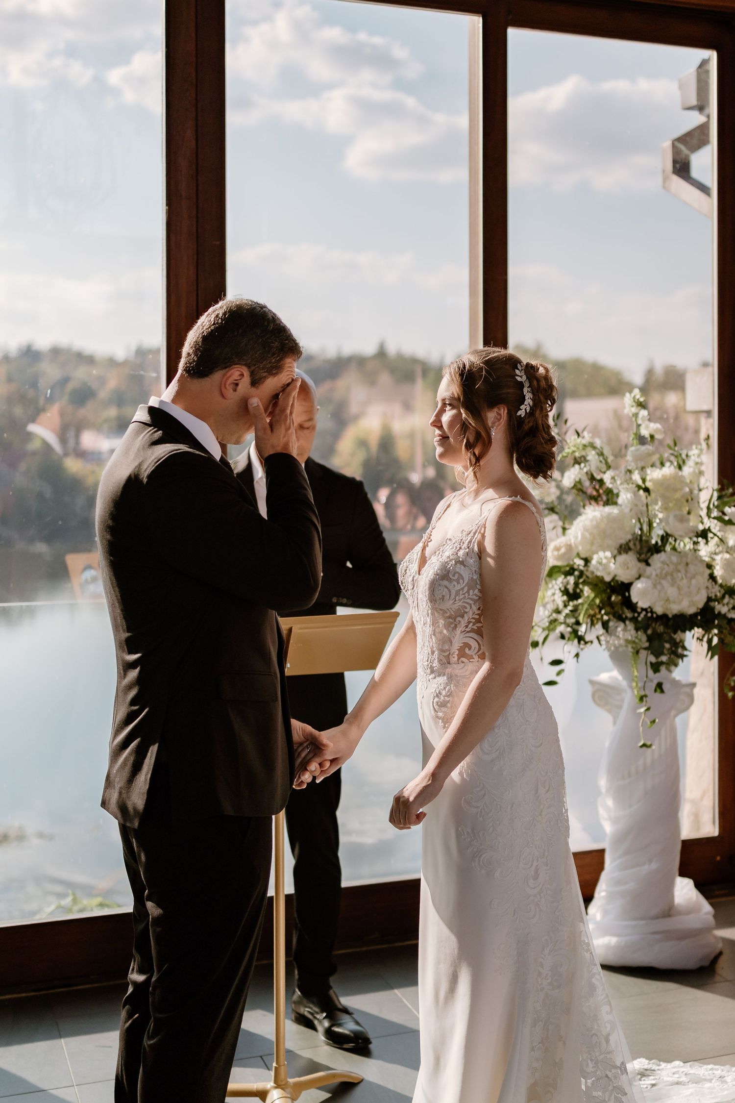 Couple exchanges vows during intimate wedding ceremony with waterfront views through large windows.