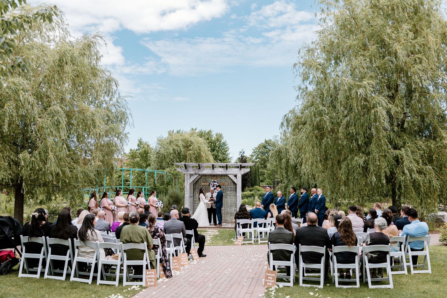Outdoor wedding ceremony with guests seated on white chairs under willow trees on a sunny day.