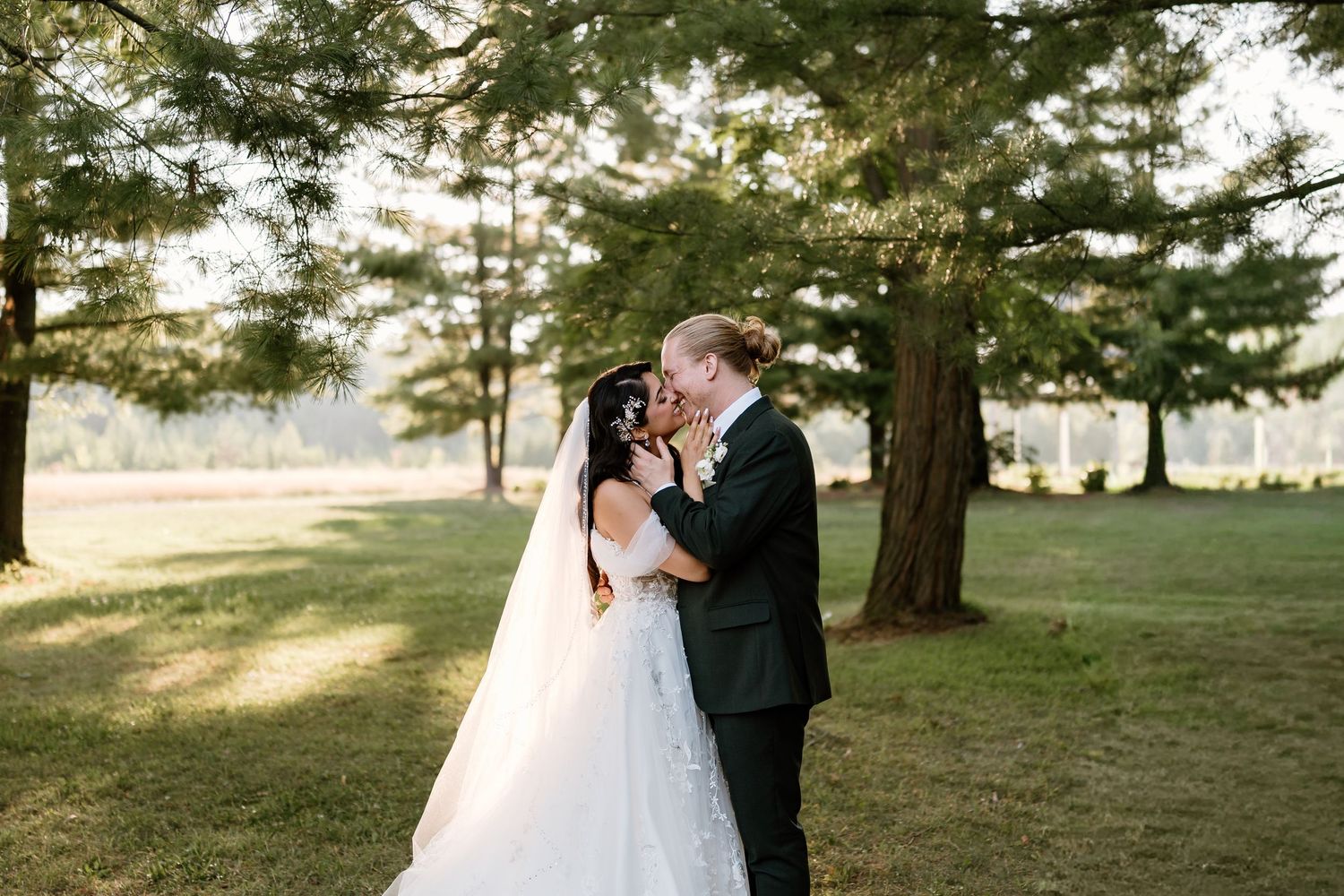 Bride and groom share a romantic kiss surrounded by tall trees in a lush green outdoor setting.