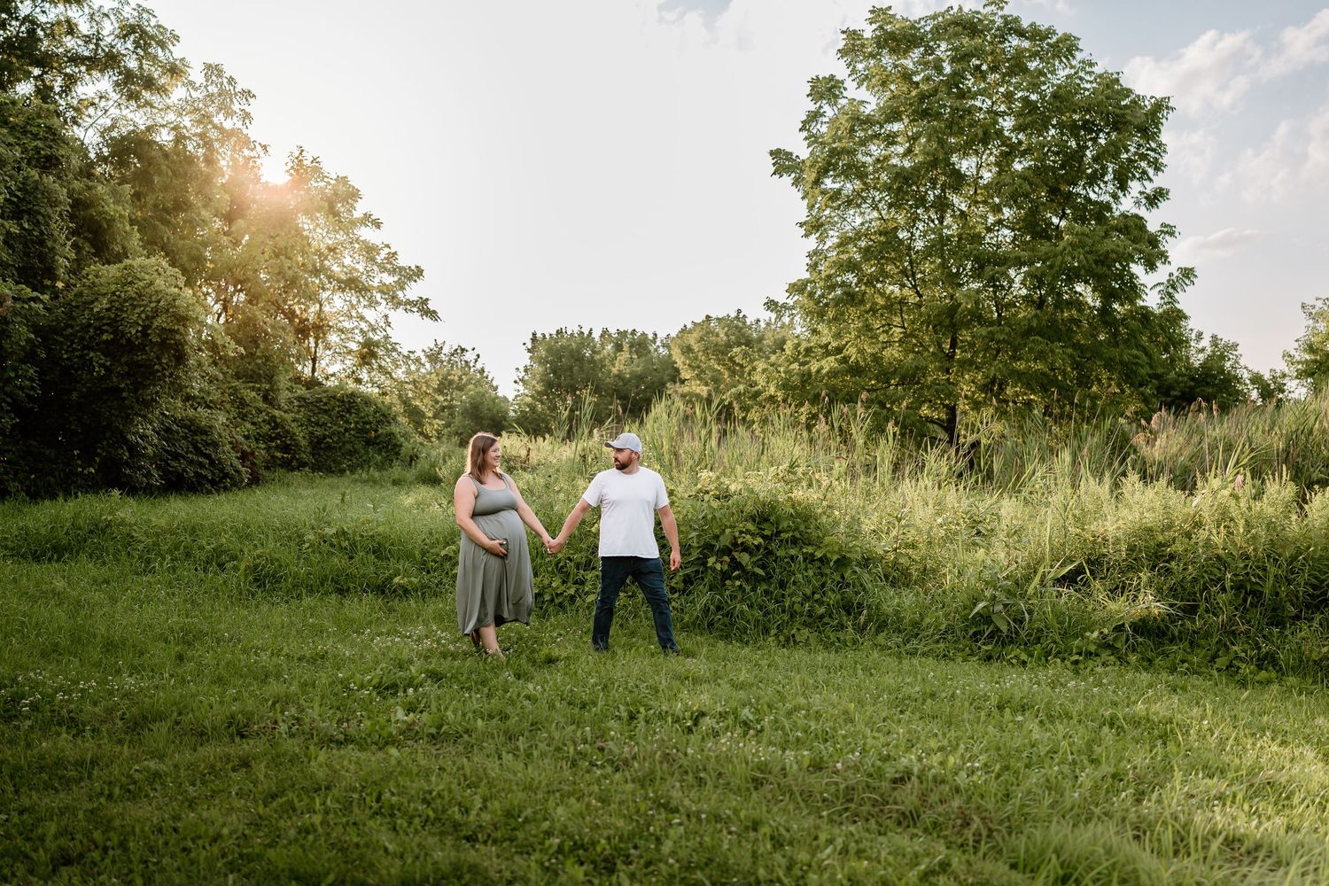 Pregnant couple holding hands in a lush green meadow during golden hour outdoor maternity photo session.
