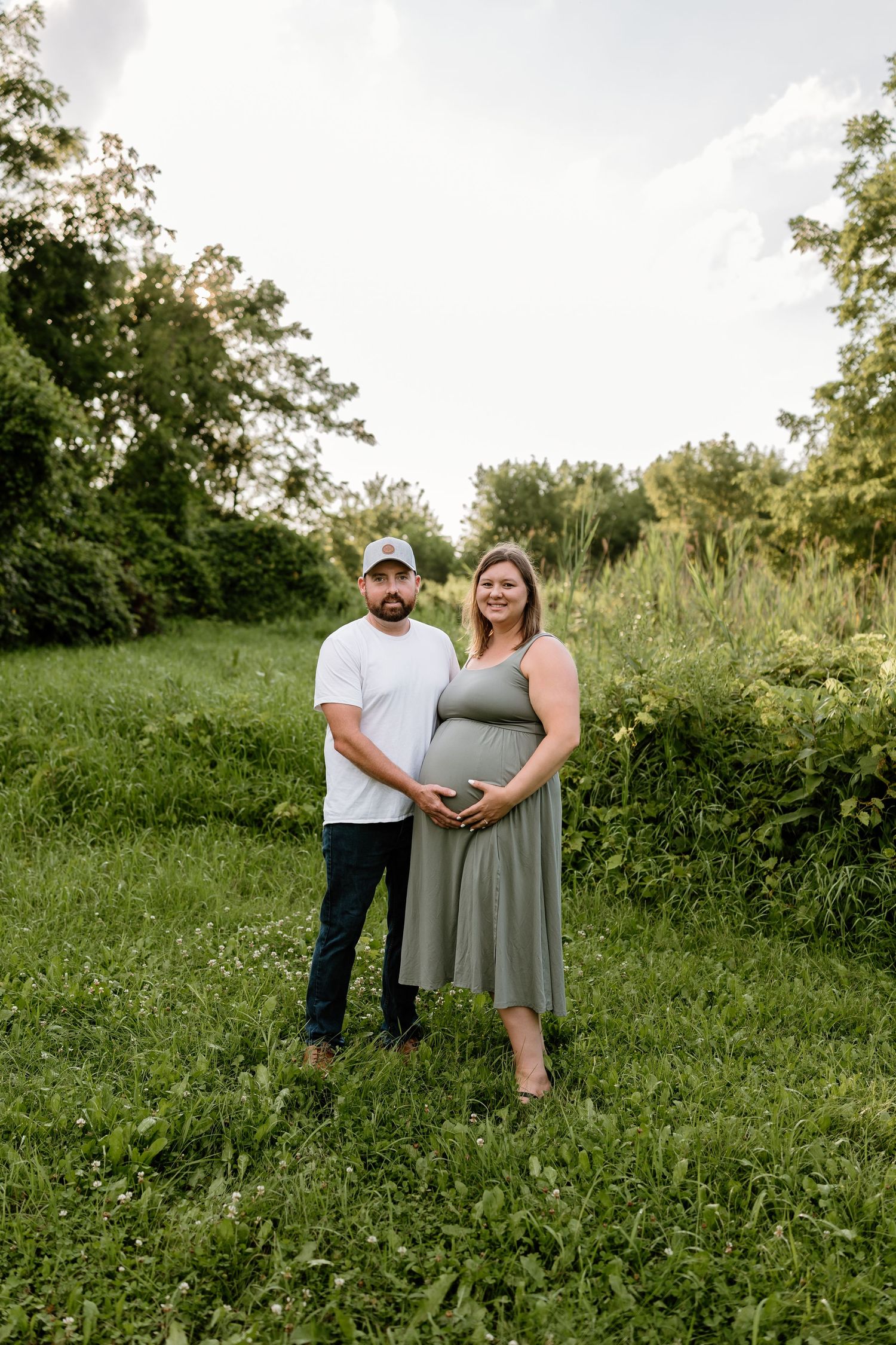 Couple poses in lush green field during outdoor maternity photo session, woman in sage dress cradling baby bump.