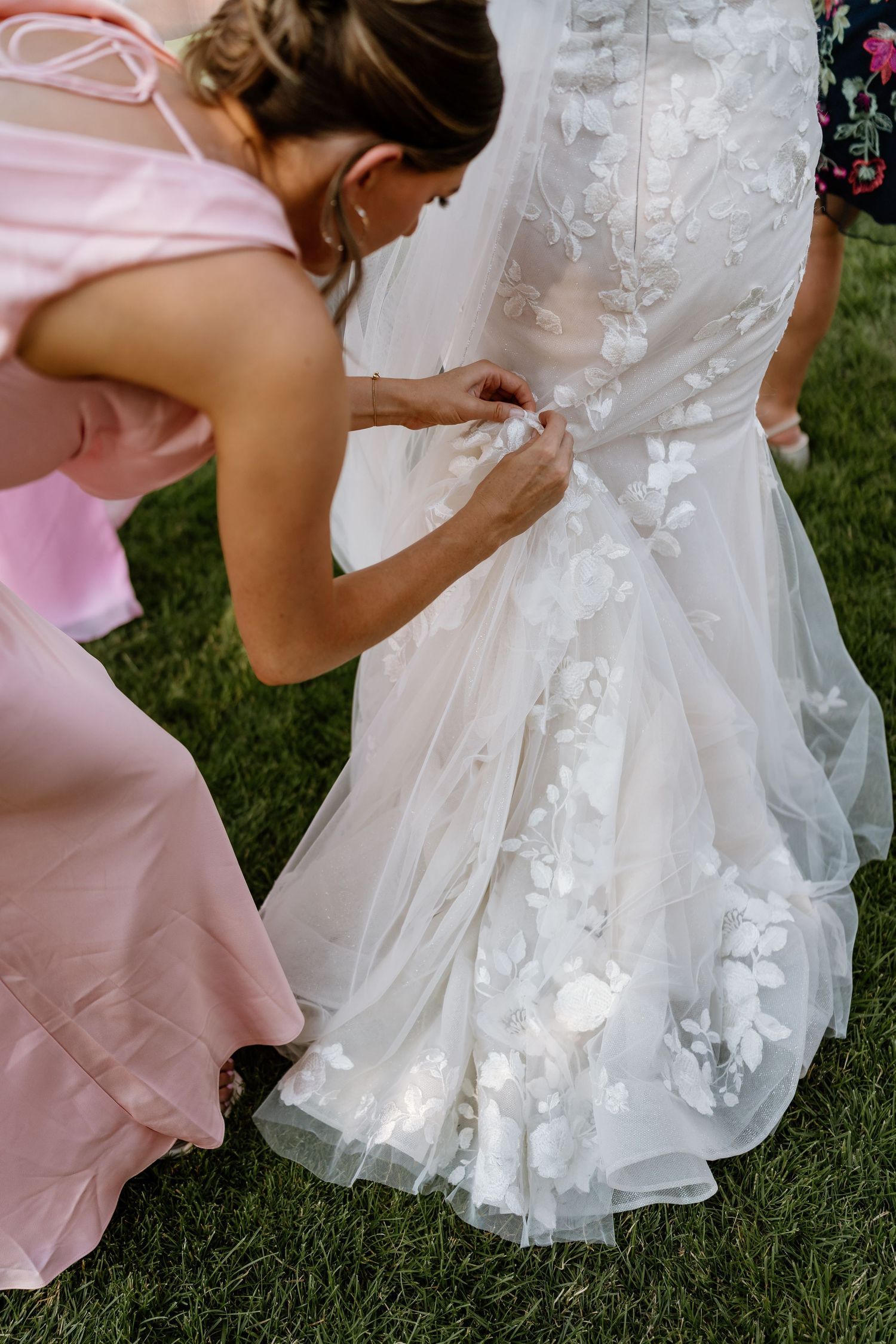 Bridesmaid in pink dress fastening buttons on brides floral lace wedding gown outdoors.