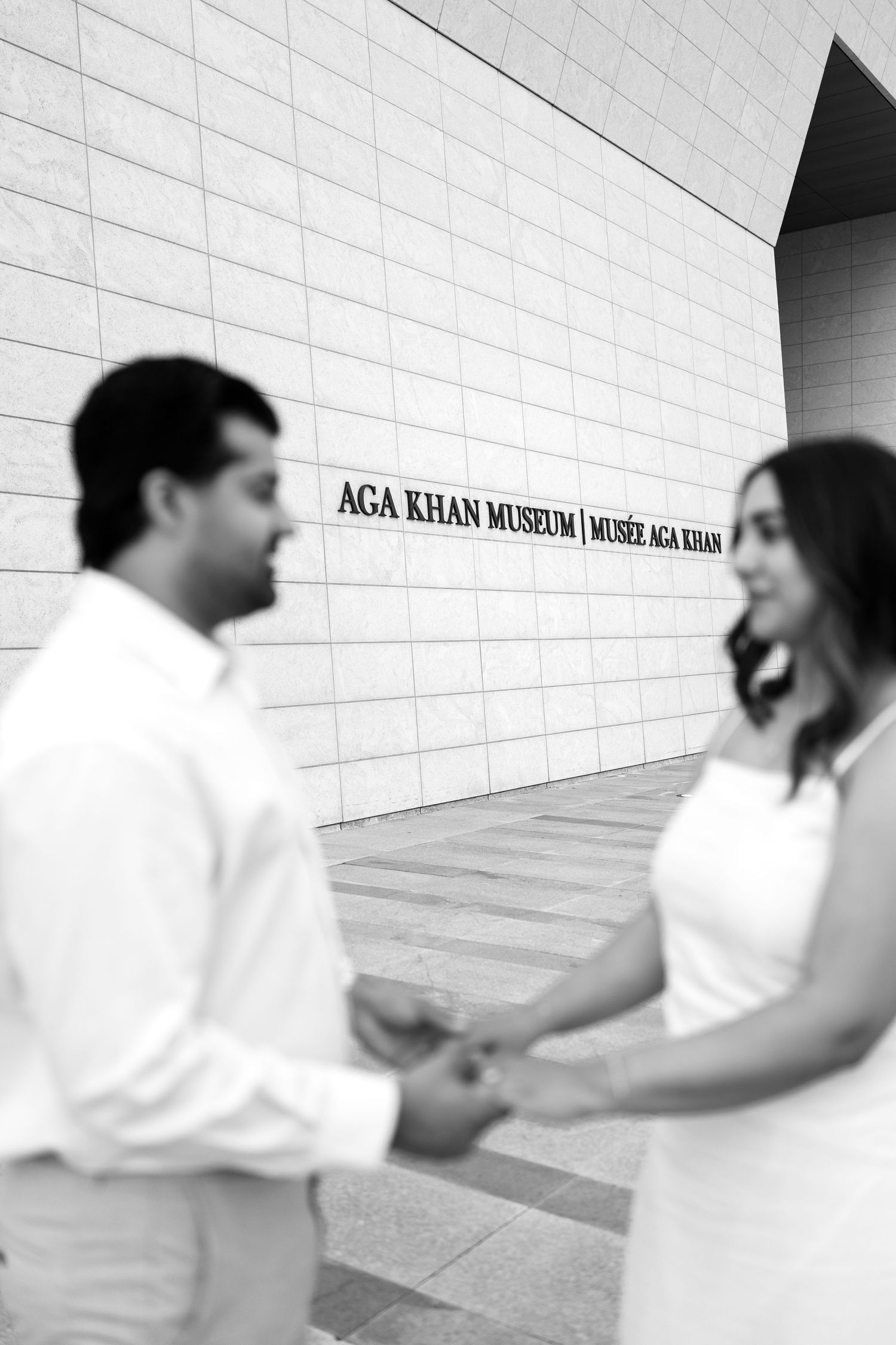Couple holds hands in front of Aga Khan Museum in black and white engagement photo.