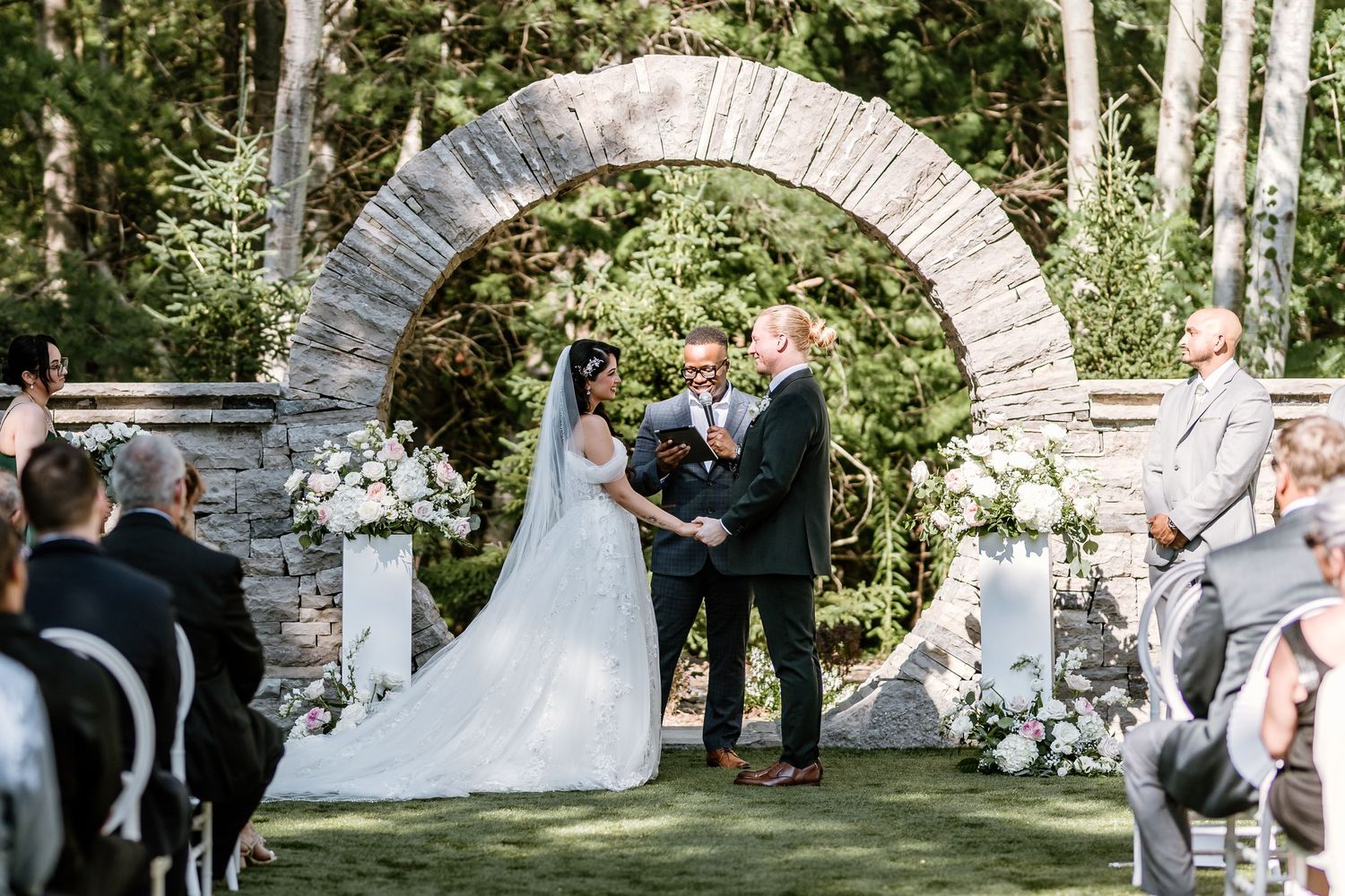 Bride and groom exchange vows under a stone arch adorned with white flowers at an outdoor wedding ceremony.